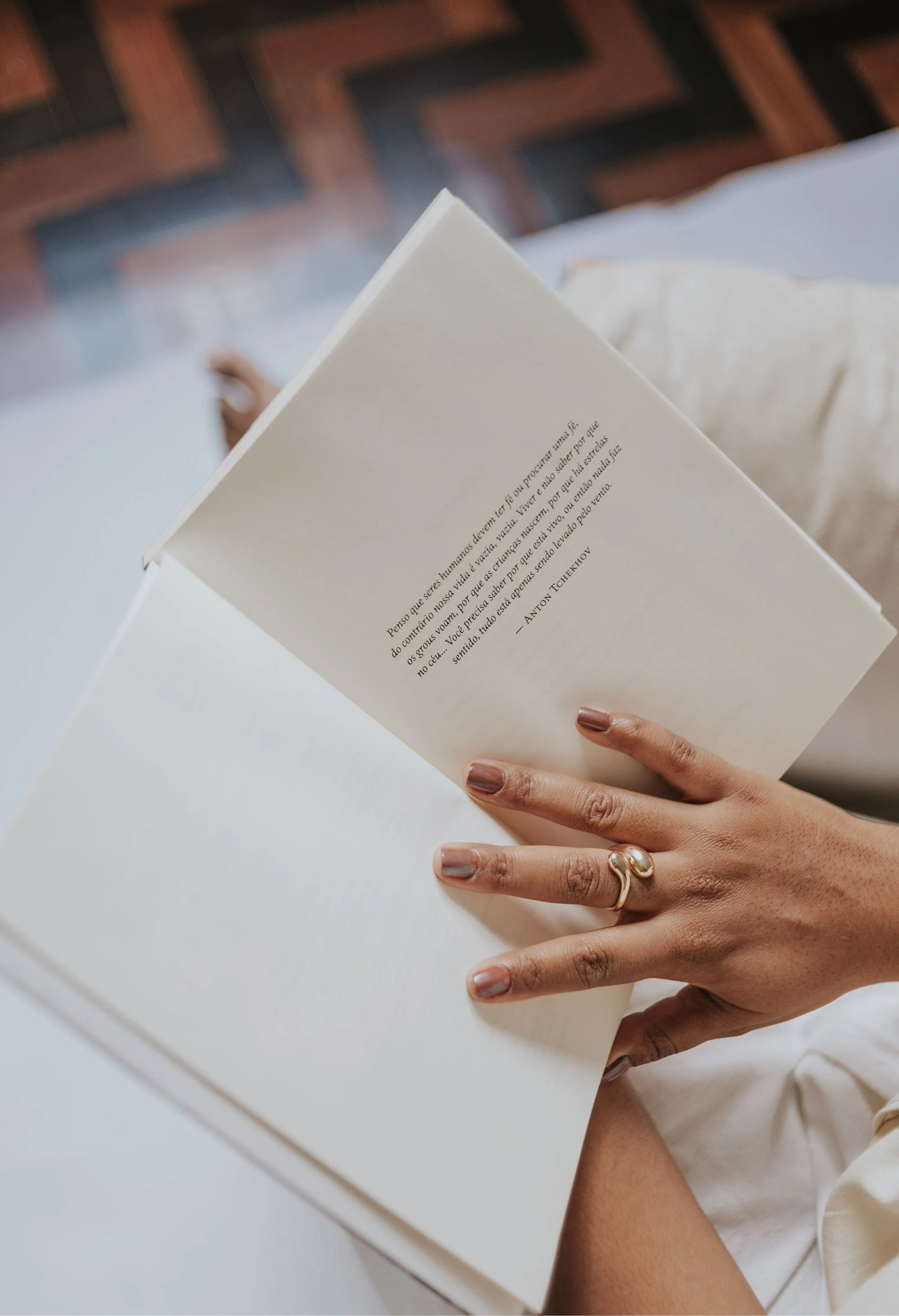 Person with painted nails and a gold ring holding a book with a quote written in Portuguese, on a white table with a blurred background.
