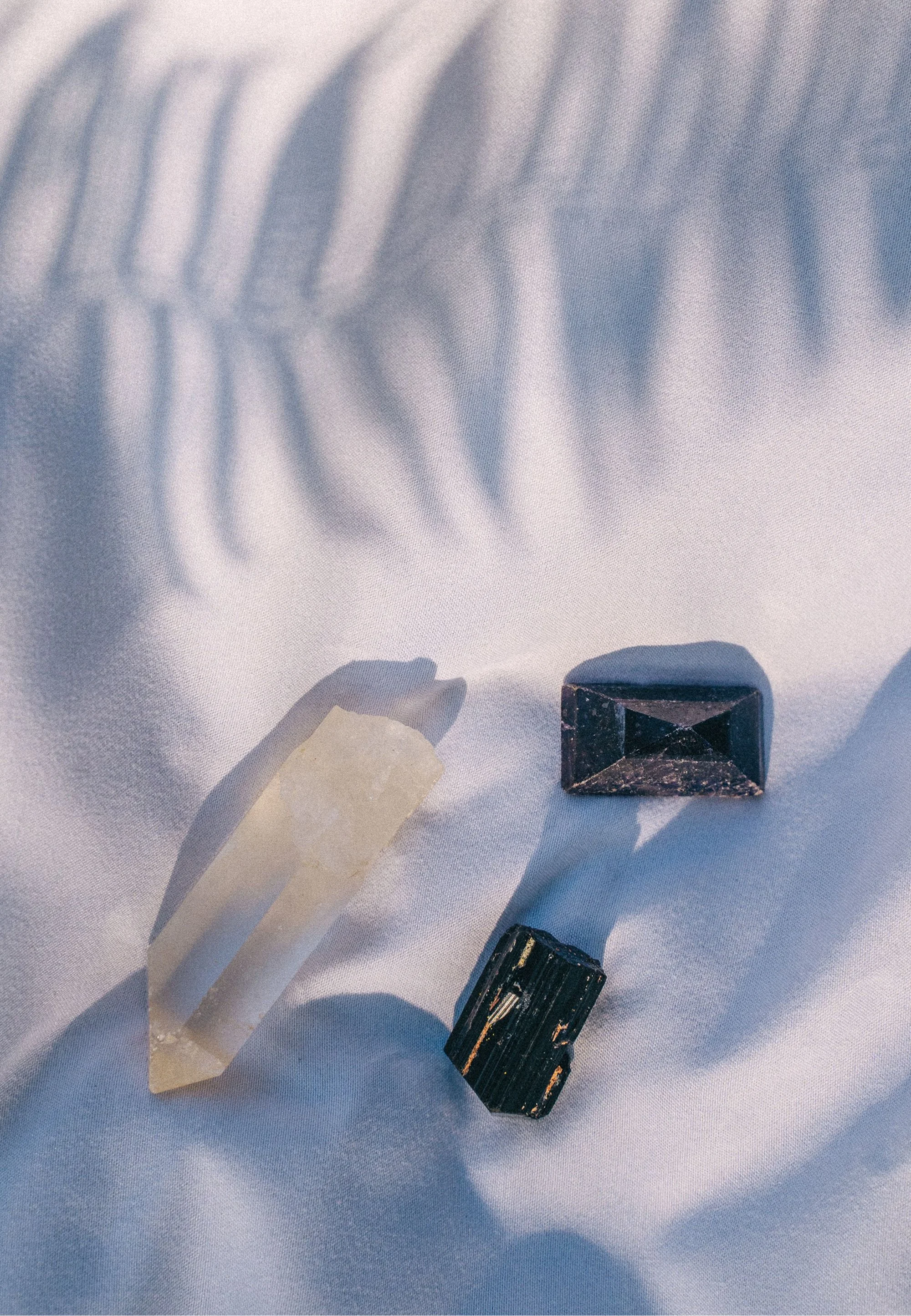 Close-up of a white fabric with shadows cast by transparent crystals, including a large clear quartz, a black tourmaline, and a black crystal.