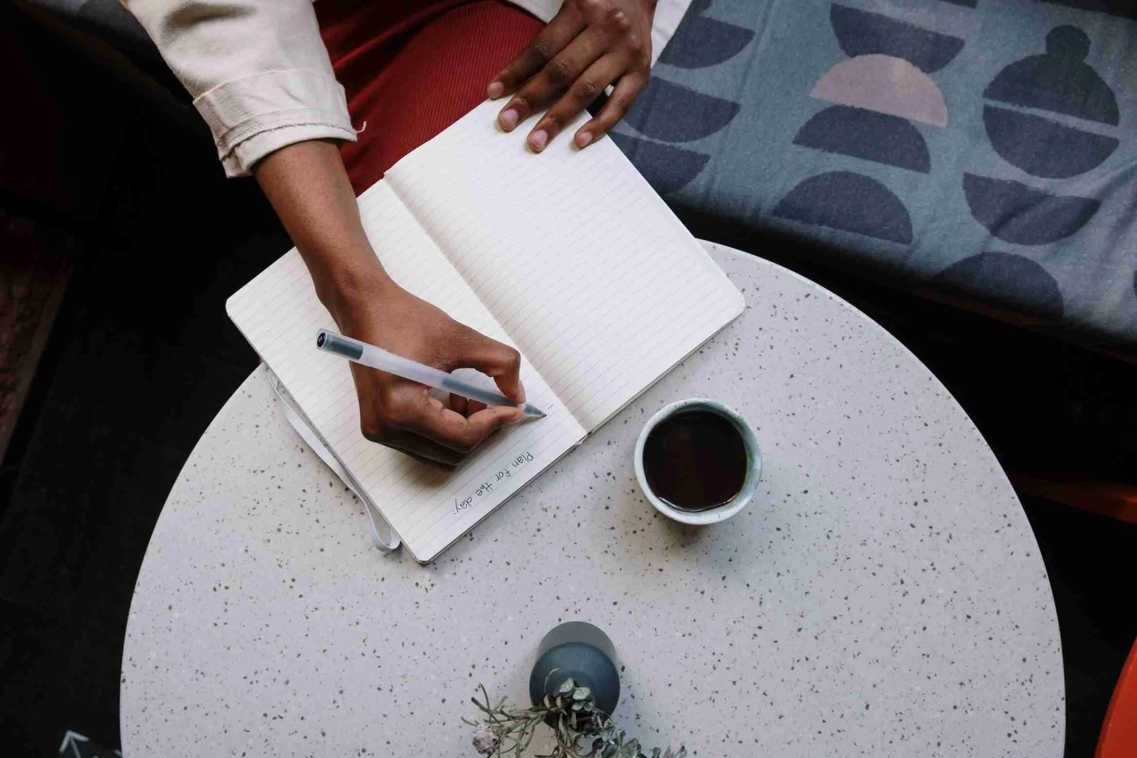 A person writing in a lined notebook with a pen, sitting at a speckled white table, with a cup of coffee and a small potted plant nearby.