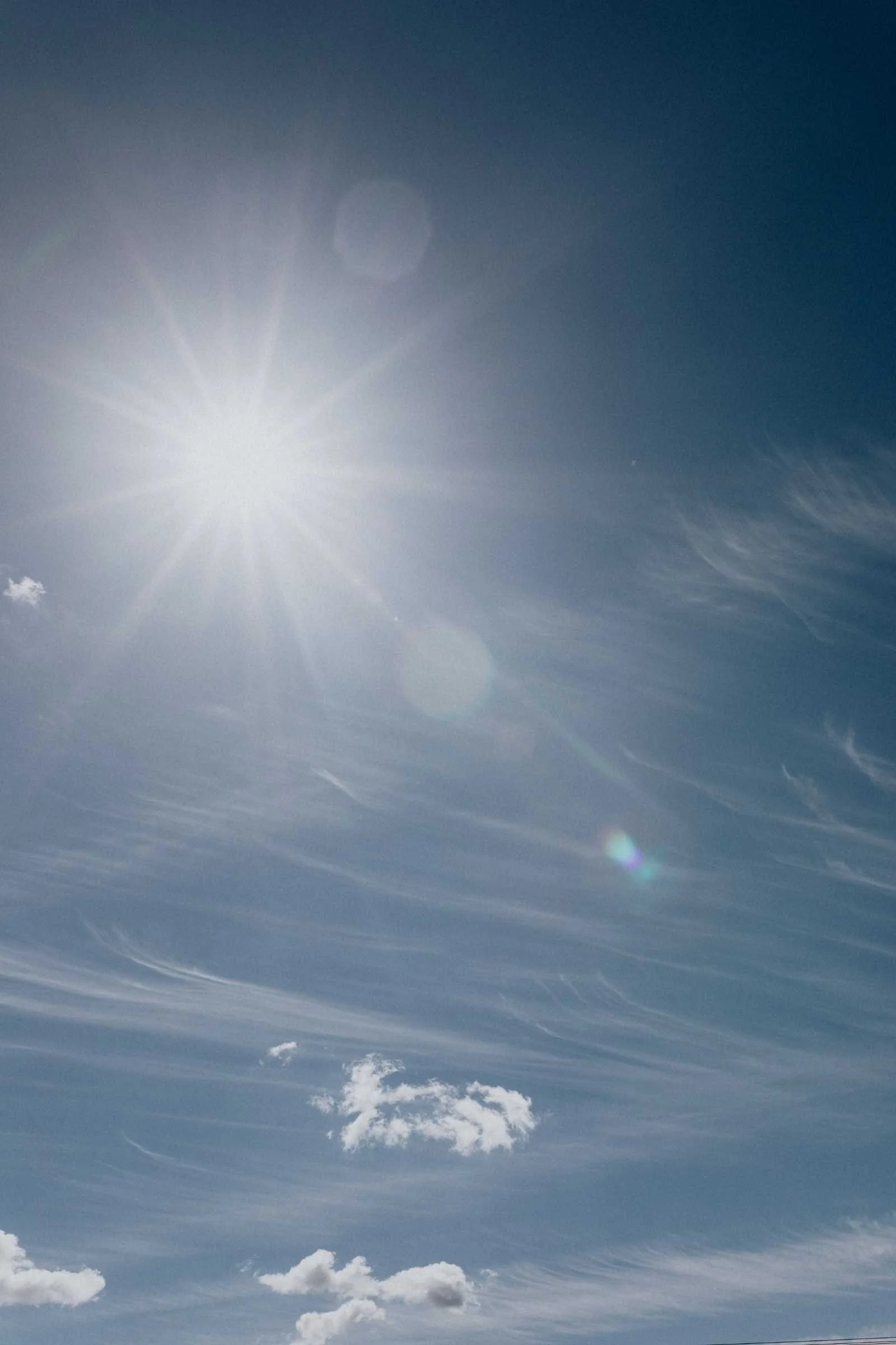 The image shows a bright sun shining in a mostly clear sky, with some white, wispy clouds scattered throughout and a slight lens flare near the center.