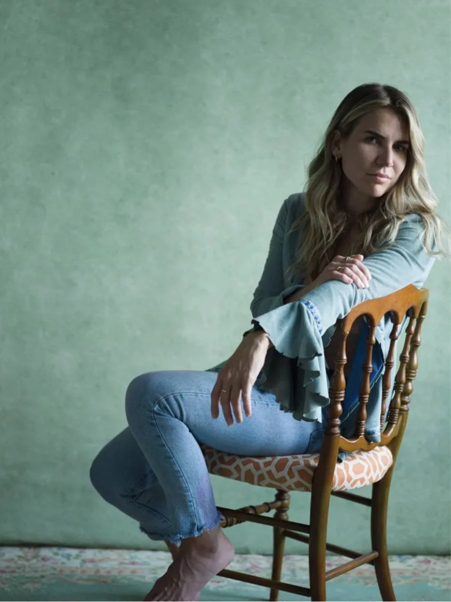 A woman with blonde hair sitting sideways on a wooden chair with a patterned cushion, wearing a light blue top and jeans, looking at the camera against a green textured background.