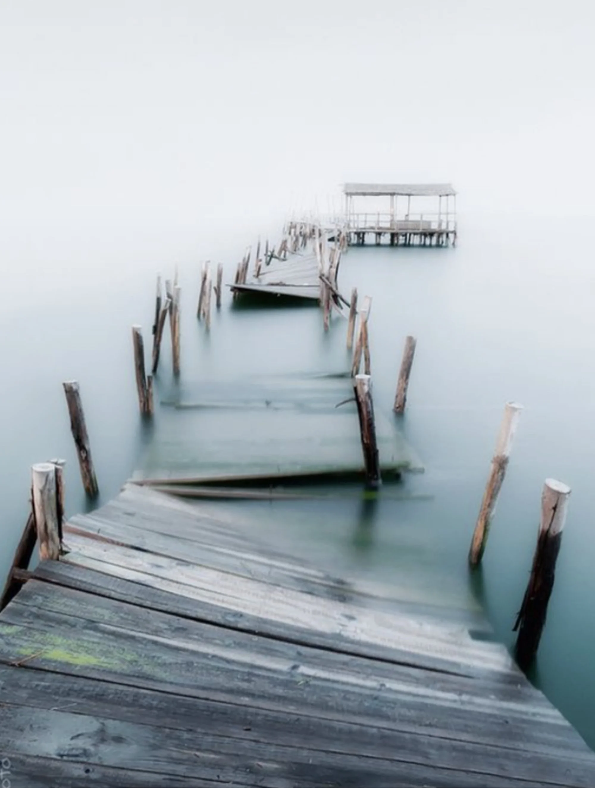 A wooden dock extending into calm water with broken and missing planks, leading to a small covered gazebo on stilts over the water. The scene appears foggy or misty.