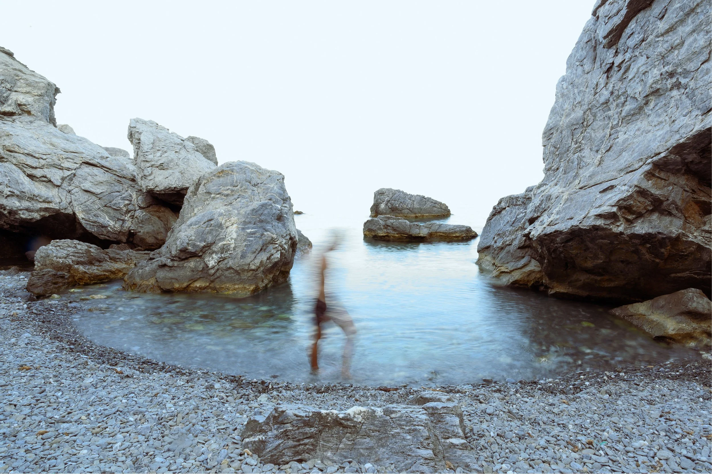 A rocky coastline with large boulders and a small body of water between cliffs under a light sky.