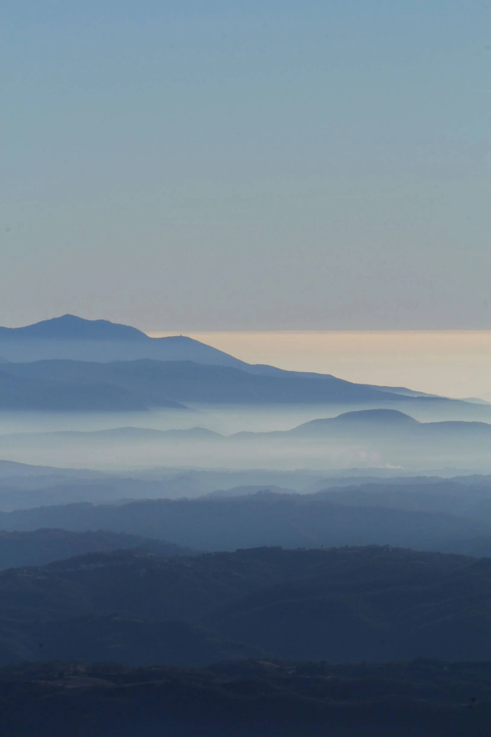 A landscape of layered mountain ranges under a pale blue sky with a hint of sunlight near the horizon.