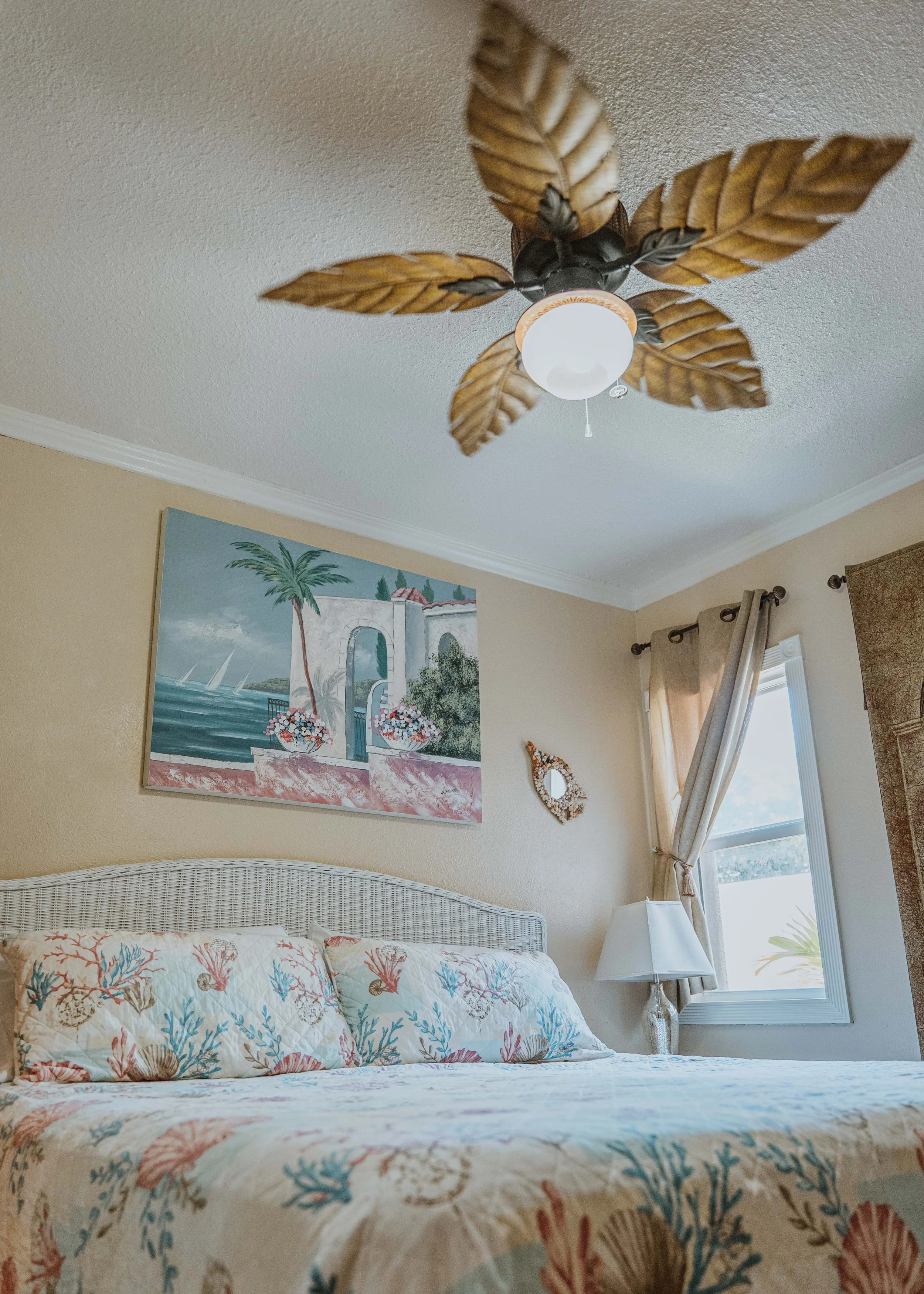 King size bed with wicker headboard and sea-themed linens underneath palm inspired ceiling fan and painting of a villa by the sea