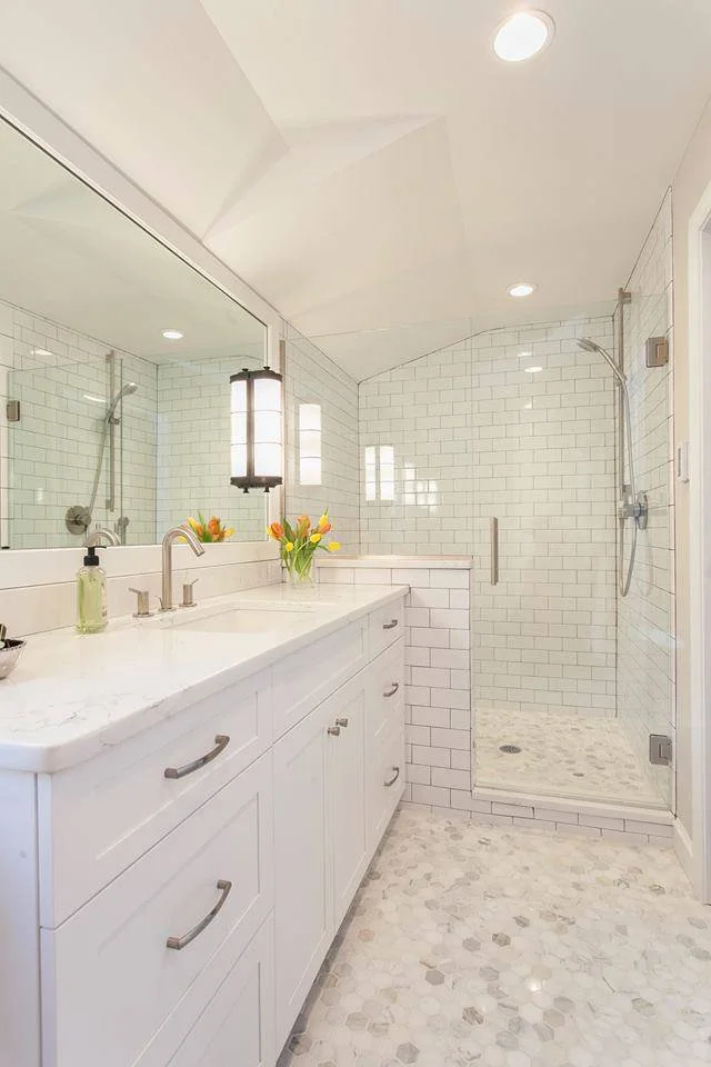 Modern white bathroom with a large mirror, white vanity with silver handles, a vase with tulips, a glass shower with white subway tiles, and pebble-style tile flooring.
