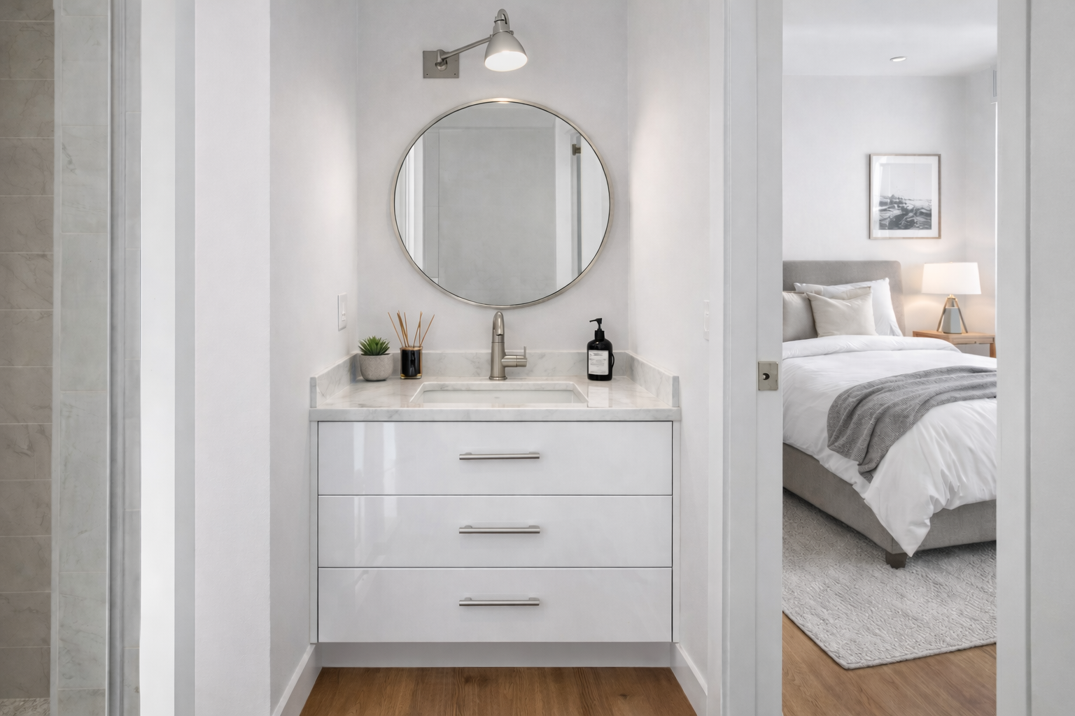 Bathroom vanity with a round mirror, a sink, a small plant, diffuser, and soap dispenser, with a bedroom visible through an open doorway.