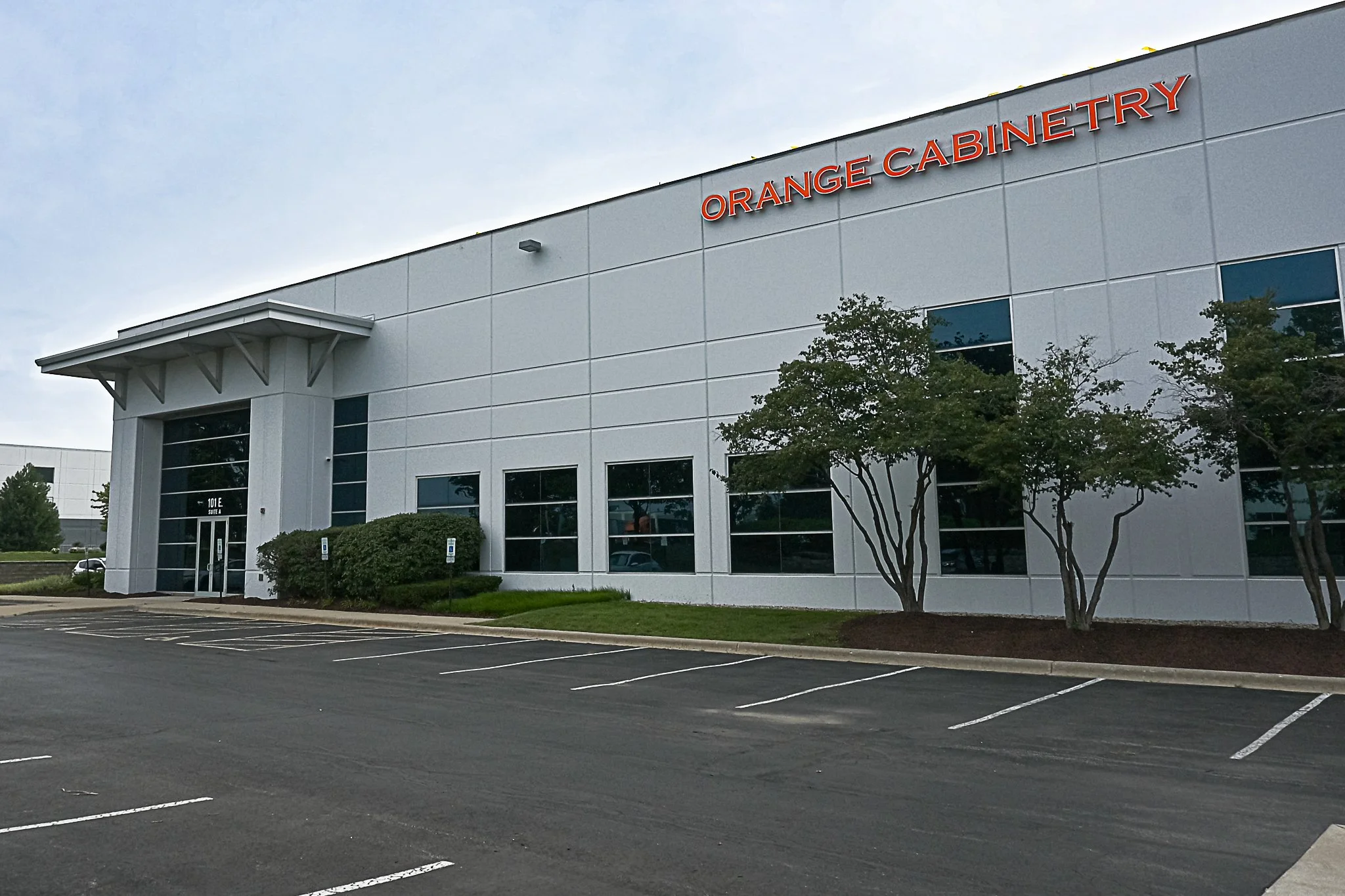 Exterior of a modern commercial building with the sign 'Orange Cabinetry', surrounded by a parking lot, green bushes, and trees.
