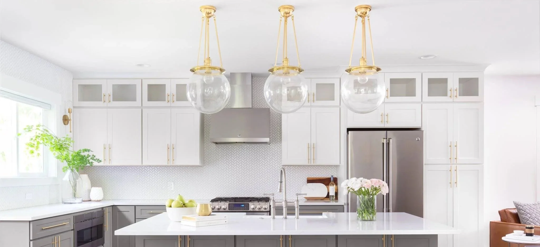 Modern kitchen with white and gray cabinetry, a large white kitchen island with a gold sink faucet, gold hardware, and three hanging globe pendant lights with gold fixtures. The kitchen features stainless steel appliances, a large refrigerator, and a