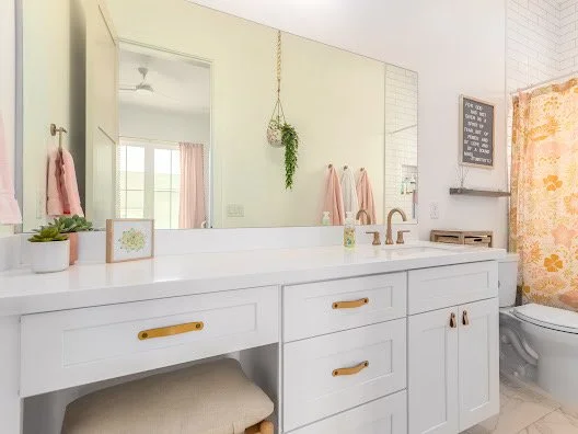 Bright bathroom with white vanity, large mirror, hanging plant, framed pictures, pink towels, and a floral shower curtain.