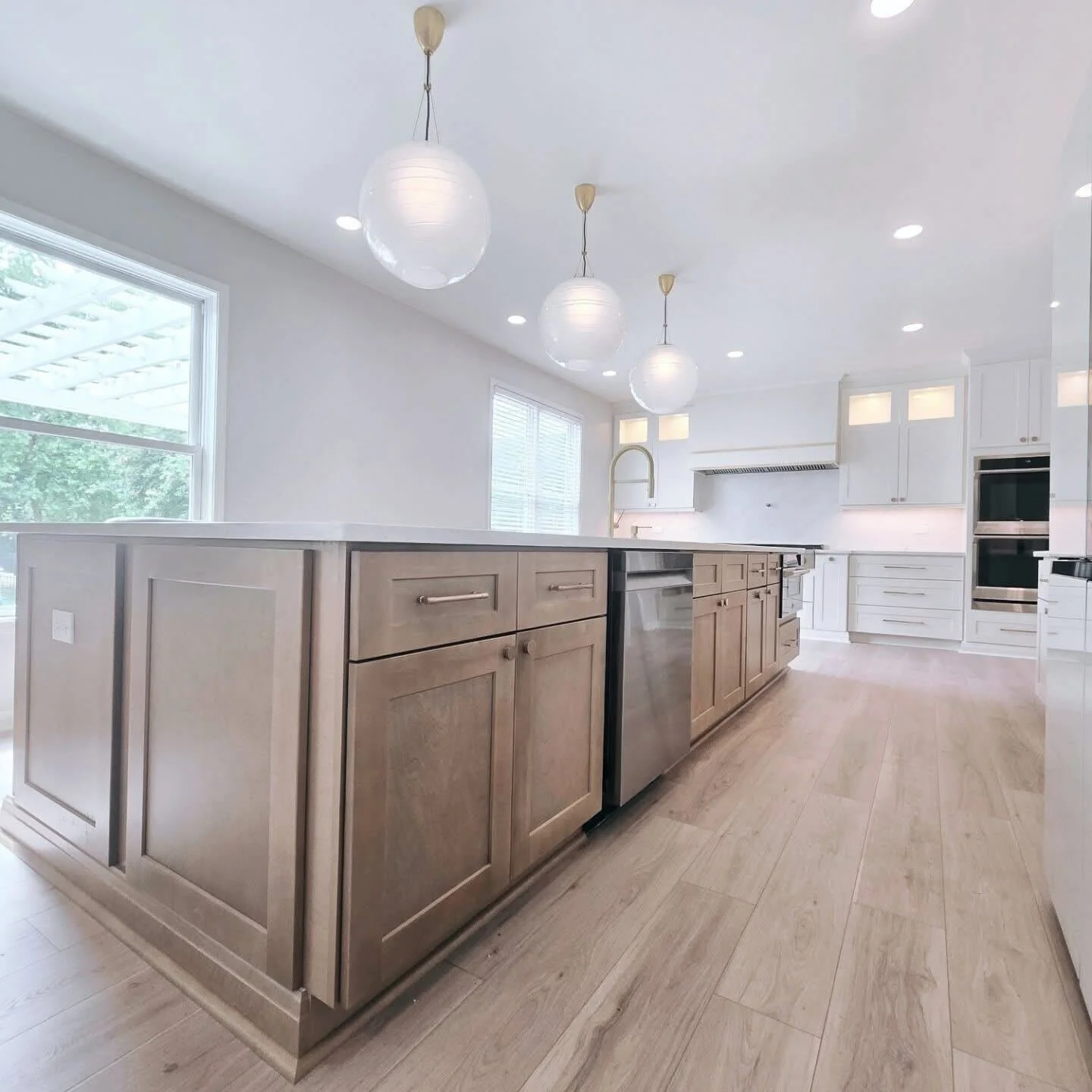 Modern kitchen with white cabinets, light wood flooring, a central island with wood panels, pendant lights, and large windows.