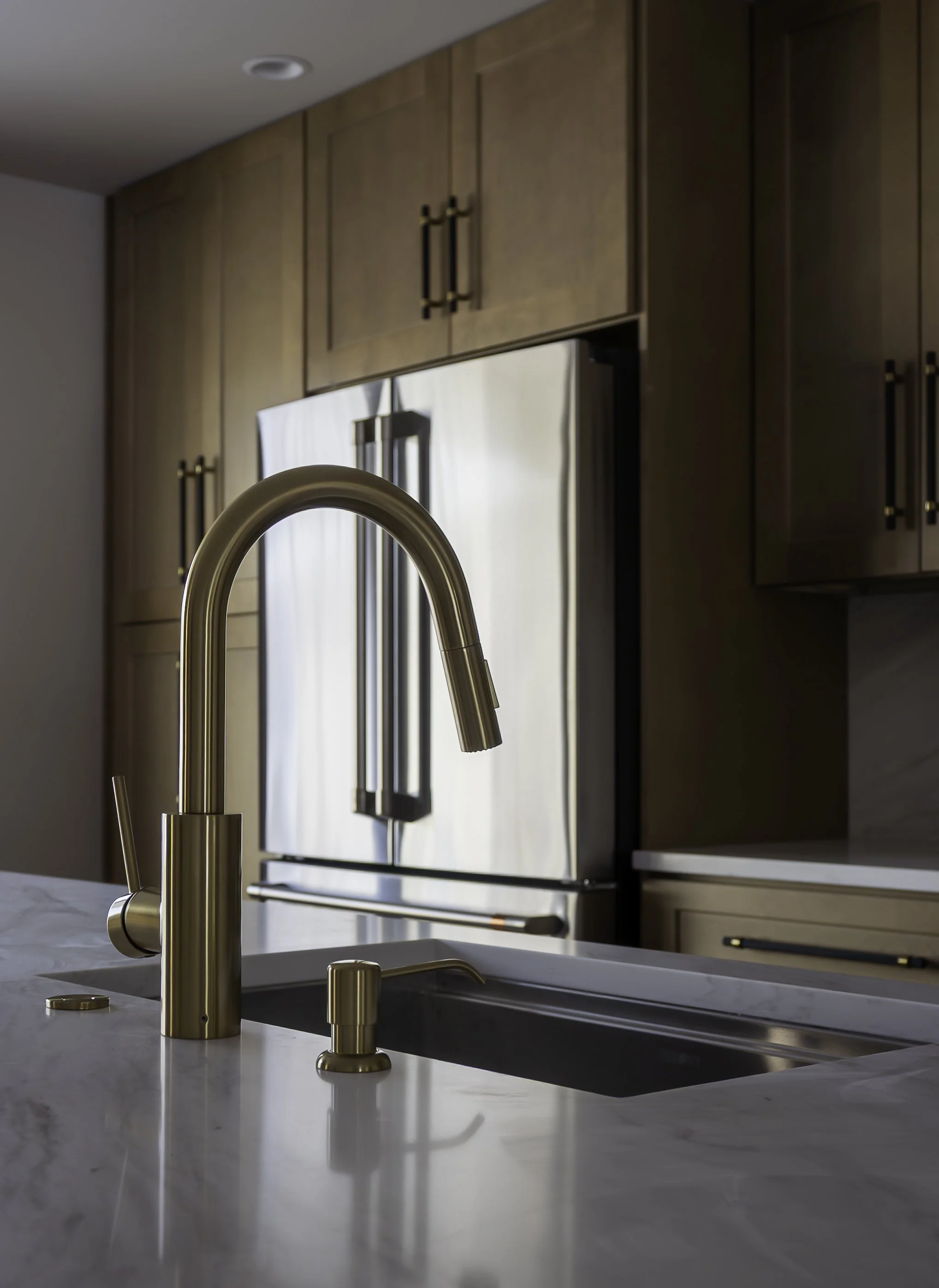 Close-up of a modern kitchen countertop with a brushed gold faucet and soap dispenser, with wooden cabinets and stainless steel refrigerator in the background.