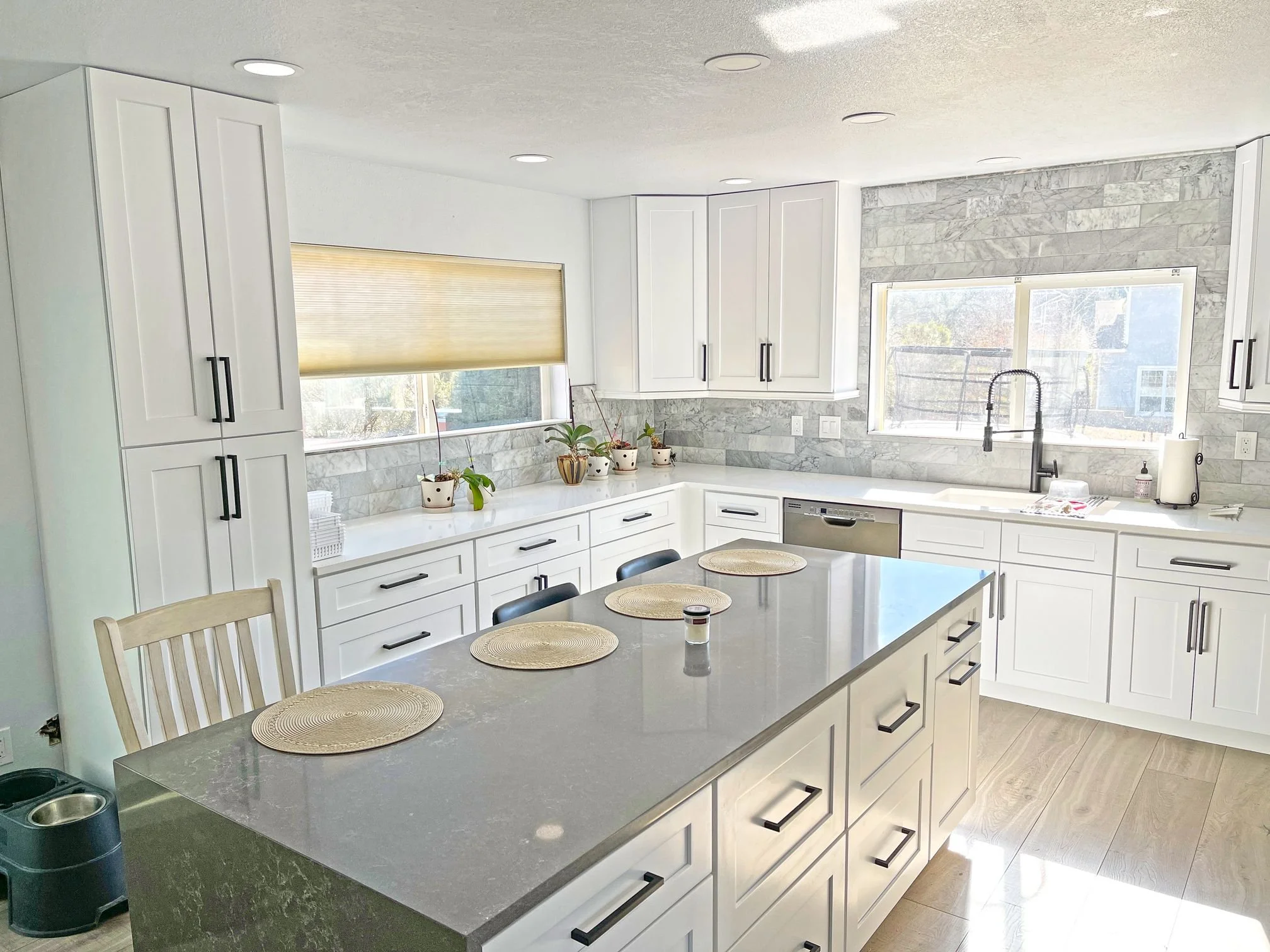Modern kitchen with white cabinets, grey countertops, and a central island. There are potted plants on the counter, and a window above the sink with a view outside. The kitchen features recessed lighting and a wood floor.