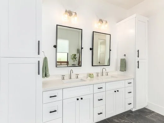 A modern white bathroom vanity with two sinks, two mirrors, and storage cabinets, illuminated by two wall-mounted light fixtures.