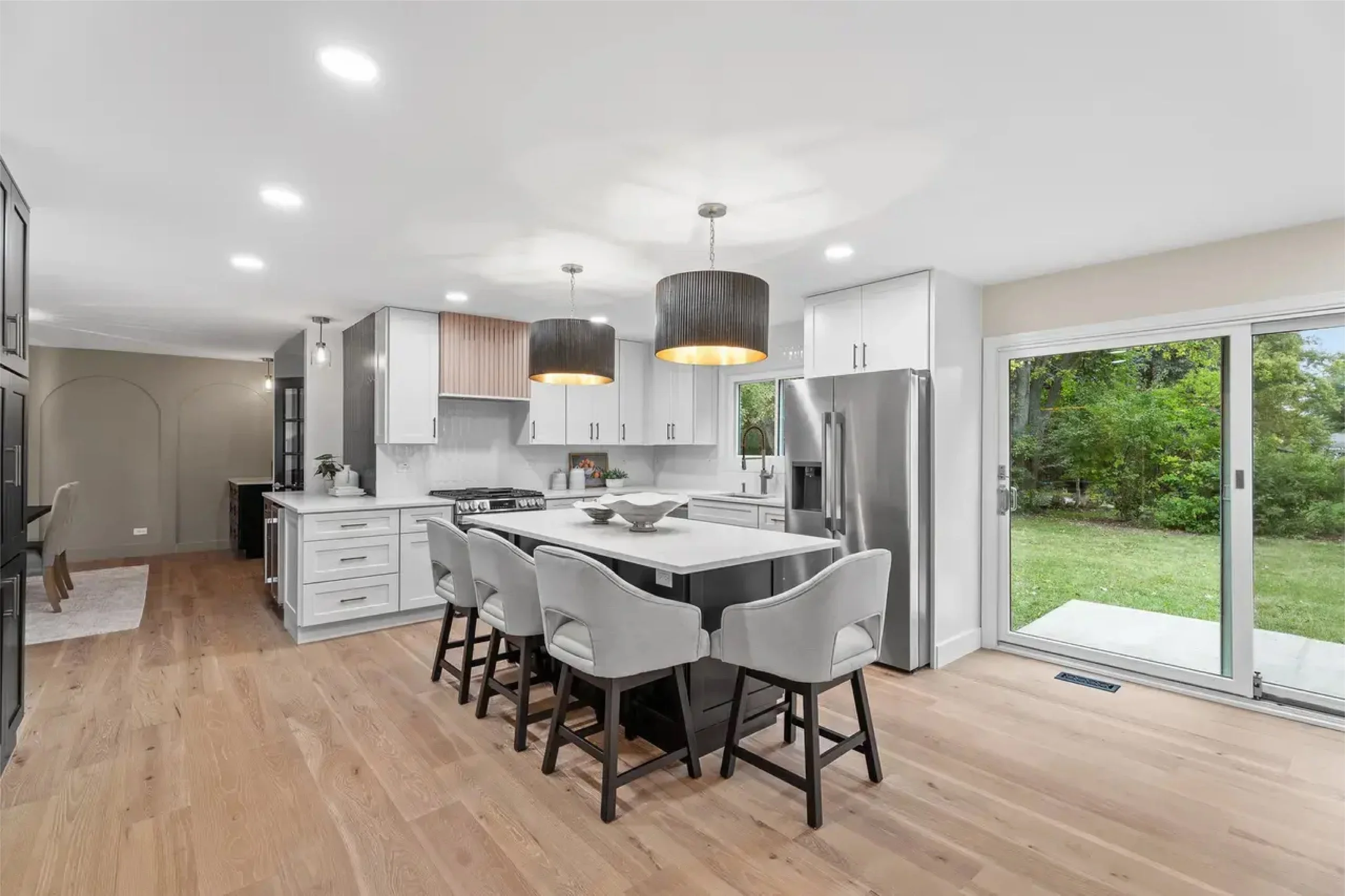 Modern kitchen with white cabinetry, stainless steel refrigerator, black pendant lights, island with bar stools, and sliding glass door opening to a green backyard.