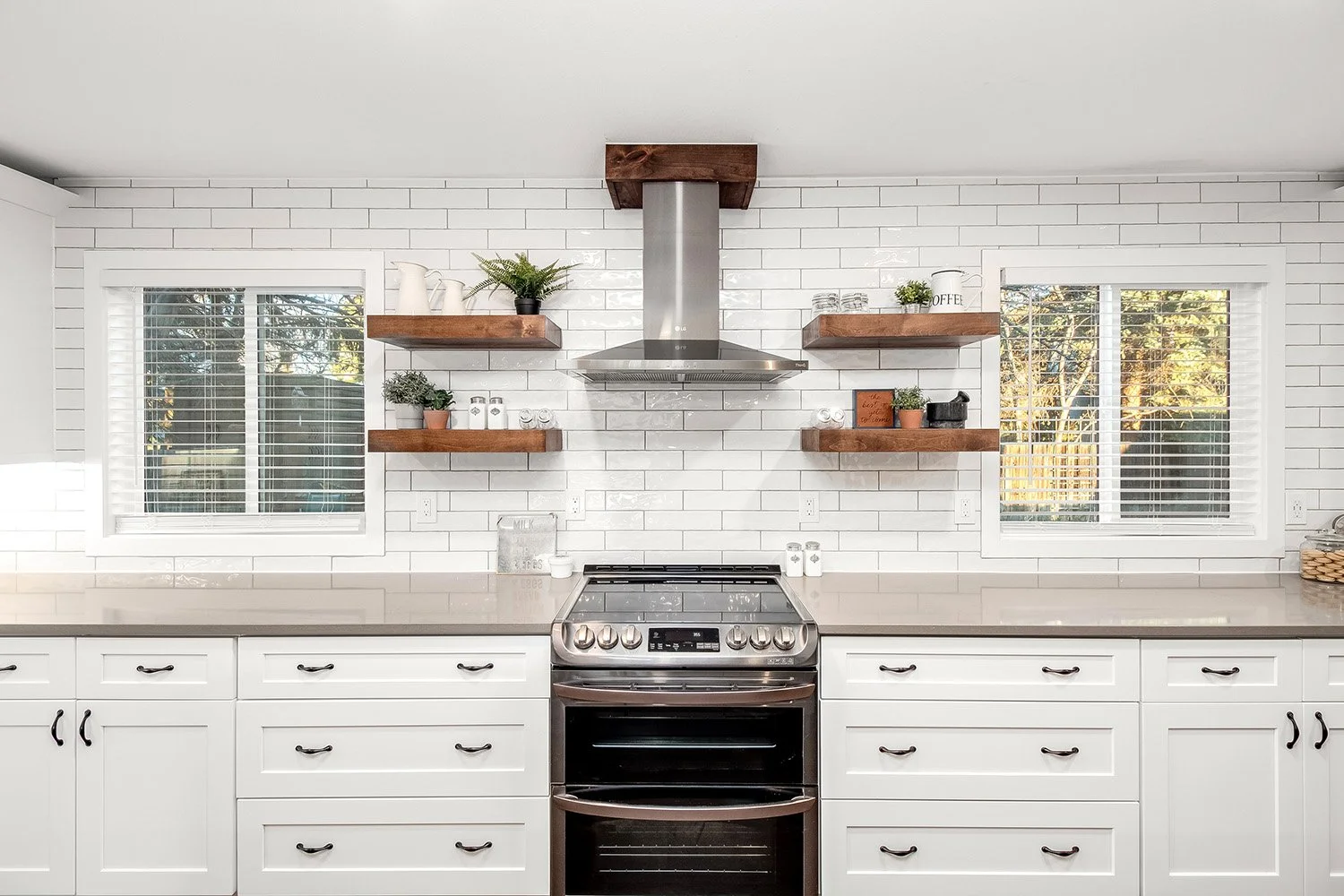 Modern kitchen with white cabinets, gray countertops, a stainless steel oven and range, two windows with white blinds, and open wooden shelves with plants and kitchen decor, white subway tile backsplash.