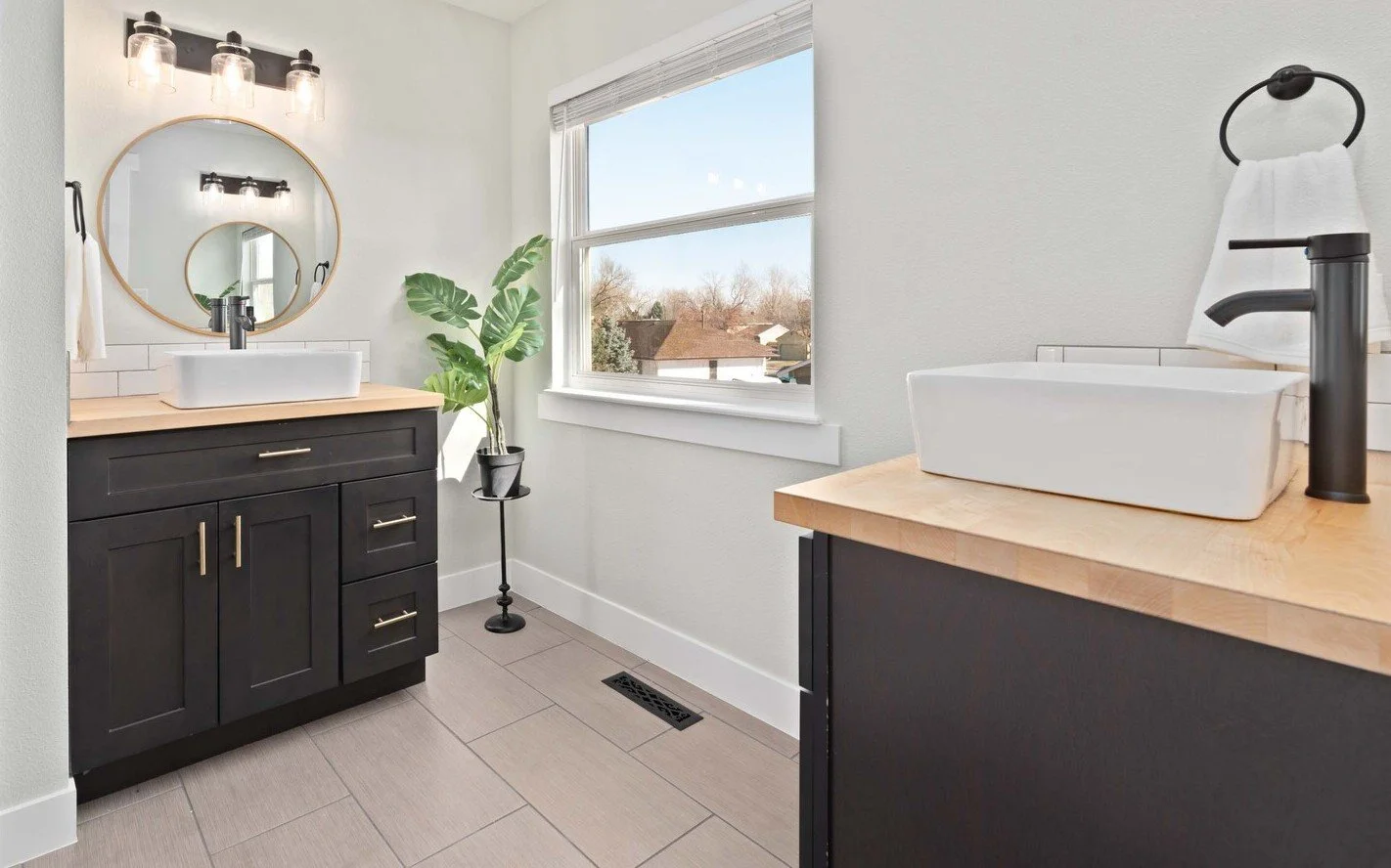Modern bathroom with white walls, black vanity with gold handles, large round mirror, vessel sink, black faucet, wooden countertop, window with a view of trees, house rooftops, and blue sky, potted plant, towel ring with a towel, light fixtures above