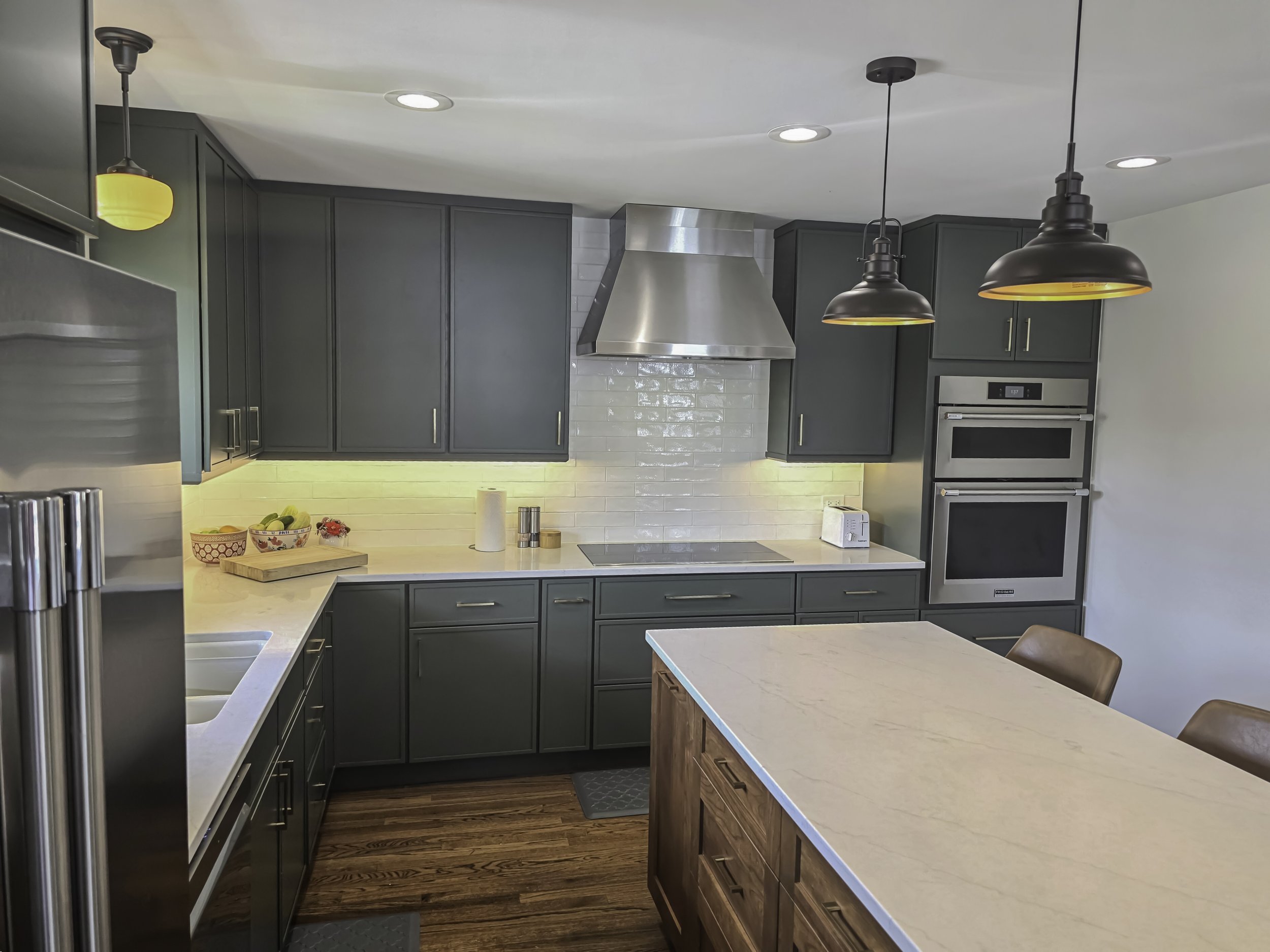 Modern kitchen with dark green cabinets, white subway tile backsplash, stainless steel appliances, and a large white island with wooden base.