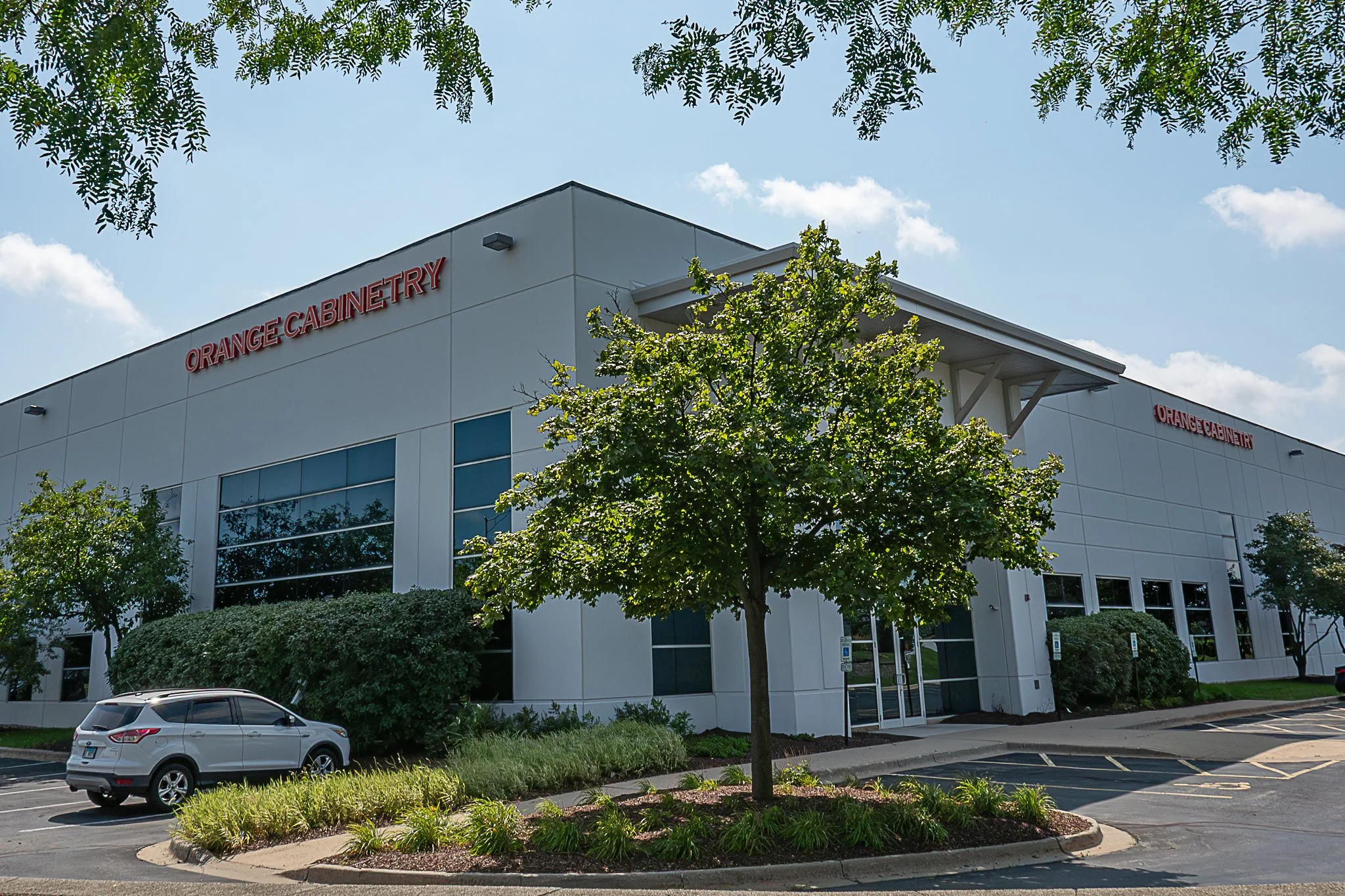 Exterior of a modern commercial building with a sign that reads 'Orange Cabinetry' in orange letters, surrounded by parking lot, trees, and bushes under a partly cloudy sky.
