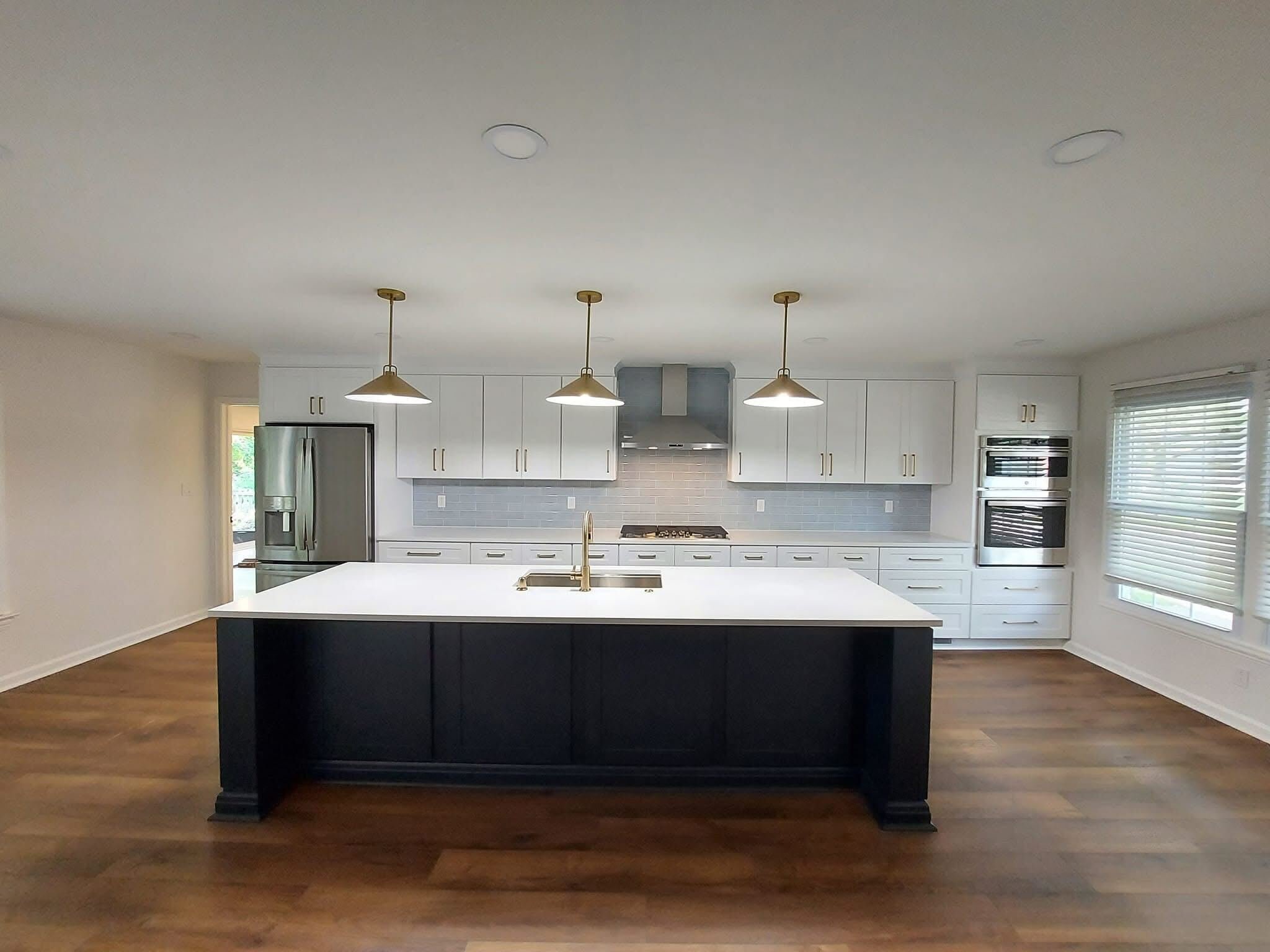 Modern kitchen with white cabinetry, gray backsplash, black island with white countertop, and hardwood flooring. Includes stainless steel refrigerator, oven, microwave, and vent hood. Three pendant lights hanging over the island.