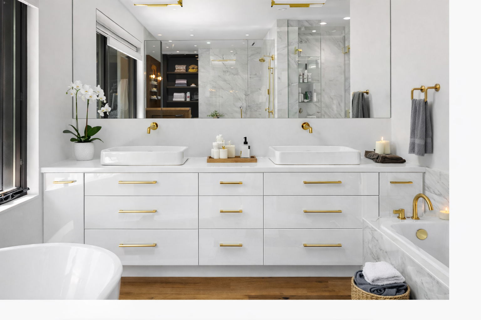 A modern white bathroom with double sinks, gold fixtures, a large mirror, and a bathtub with candles. There is a flower in a white pot, towels, and a wicker basket with folded towels.