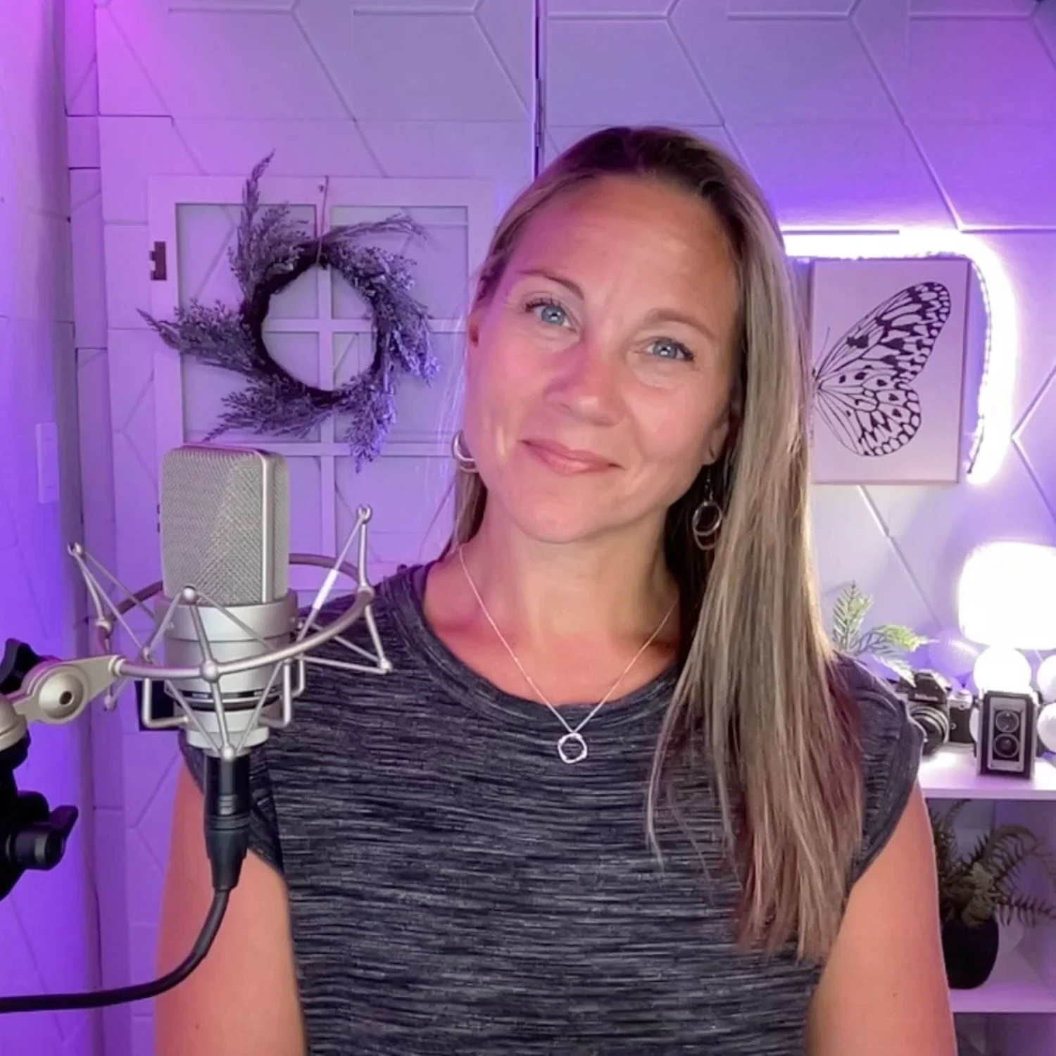 A woman with long blonde hair and blue eyes smiling at the camera, sitting in front of a professional microphone in a room with purple ambient lighting and decorative wall art, including a butterfly and a wreath of flowers.