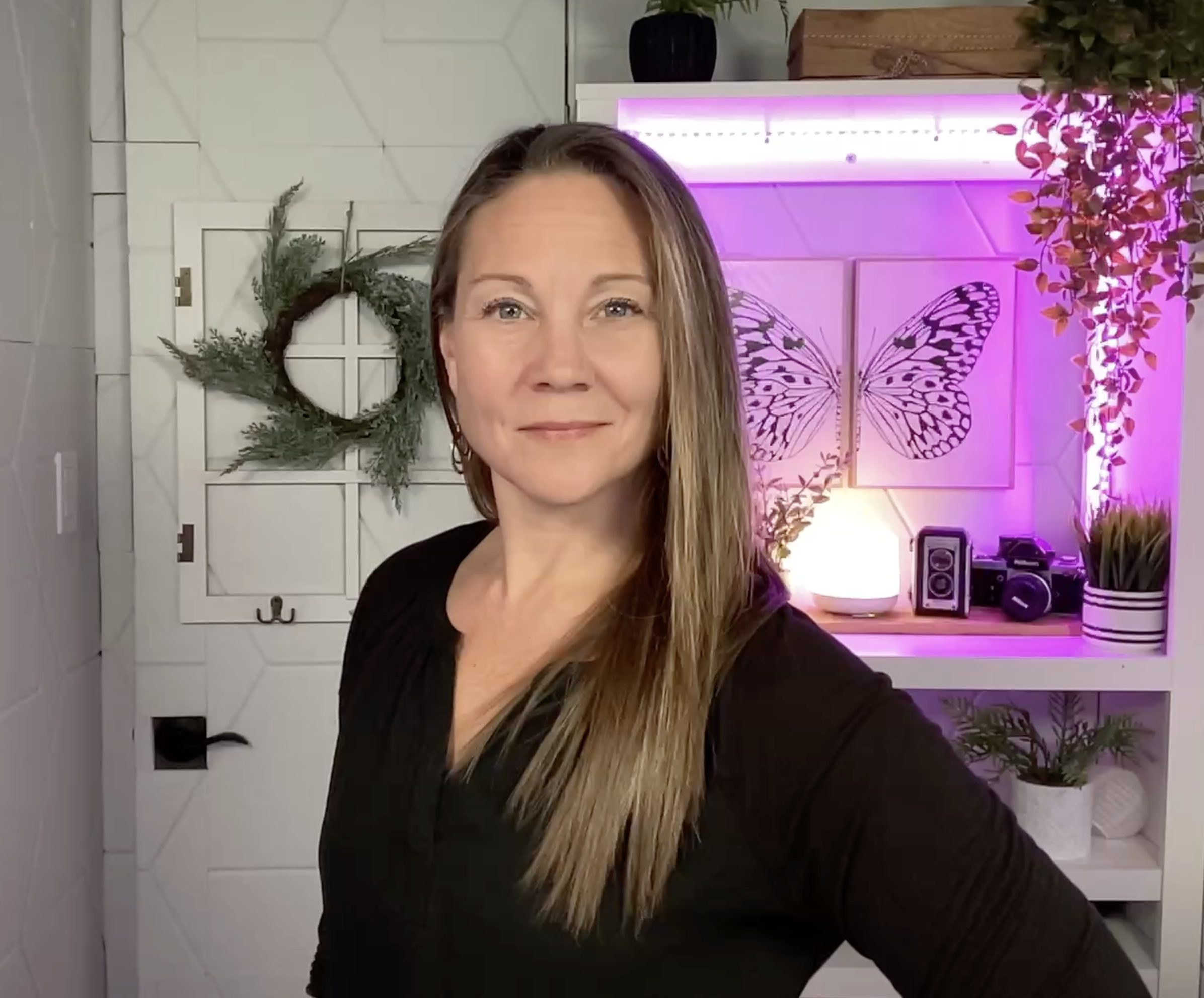 A woman with long hair smiling, sitting in a room with white tiles and plants. The background features a wall with a butterfly image and pink lighting, with decorative items like a radio, speaker, and plant pots.