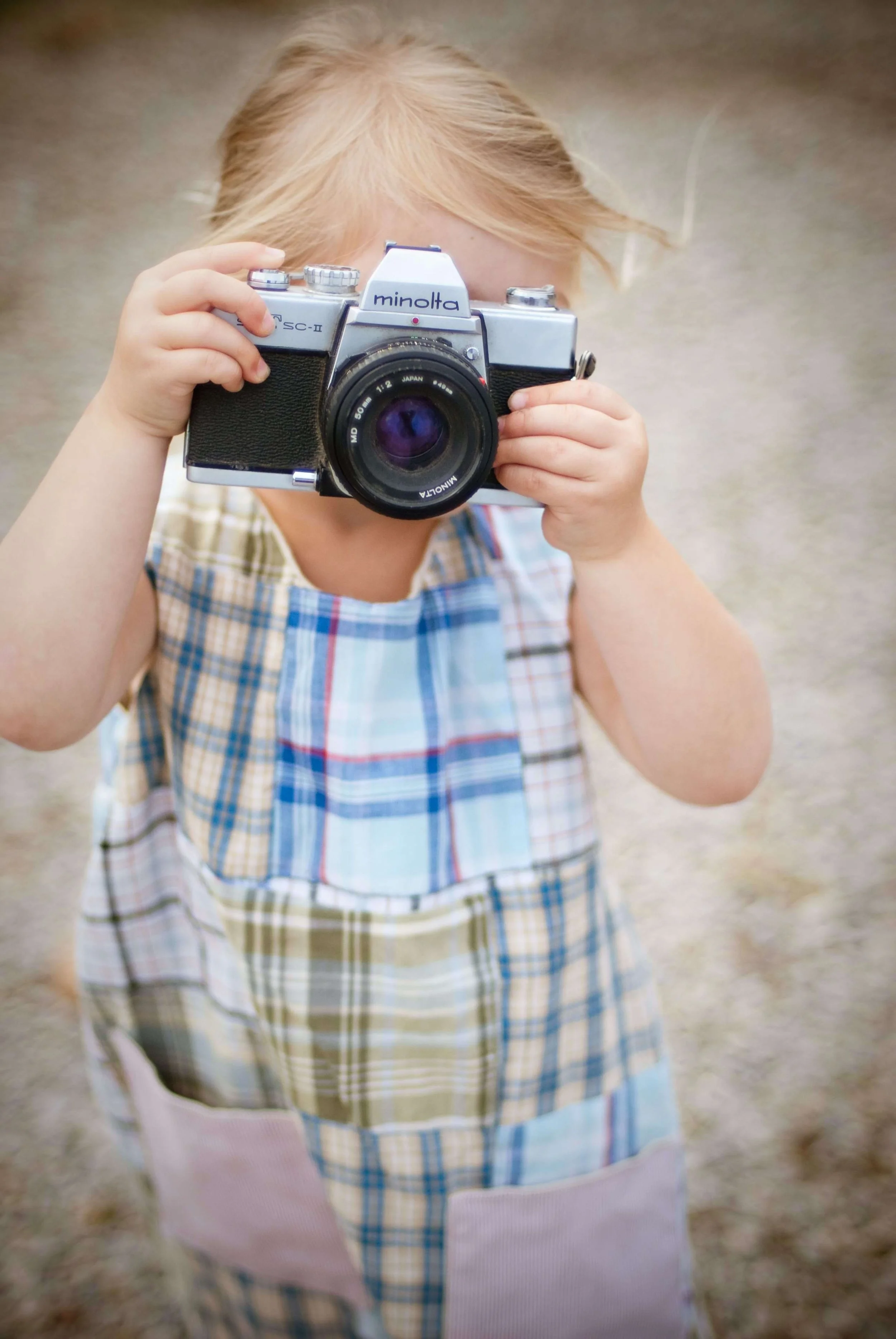 A young girl with blonde hair holding a vintage Minolta camera up to her face, standing outdoors on a neutral background.
