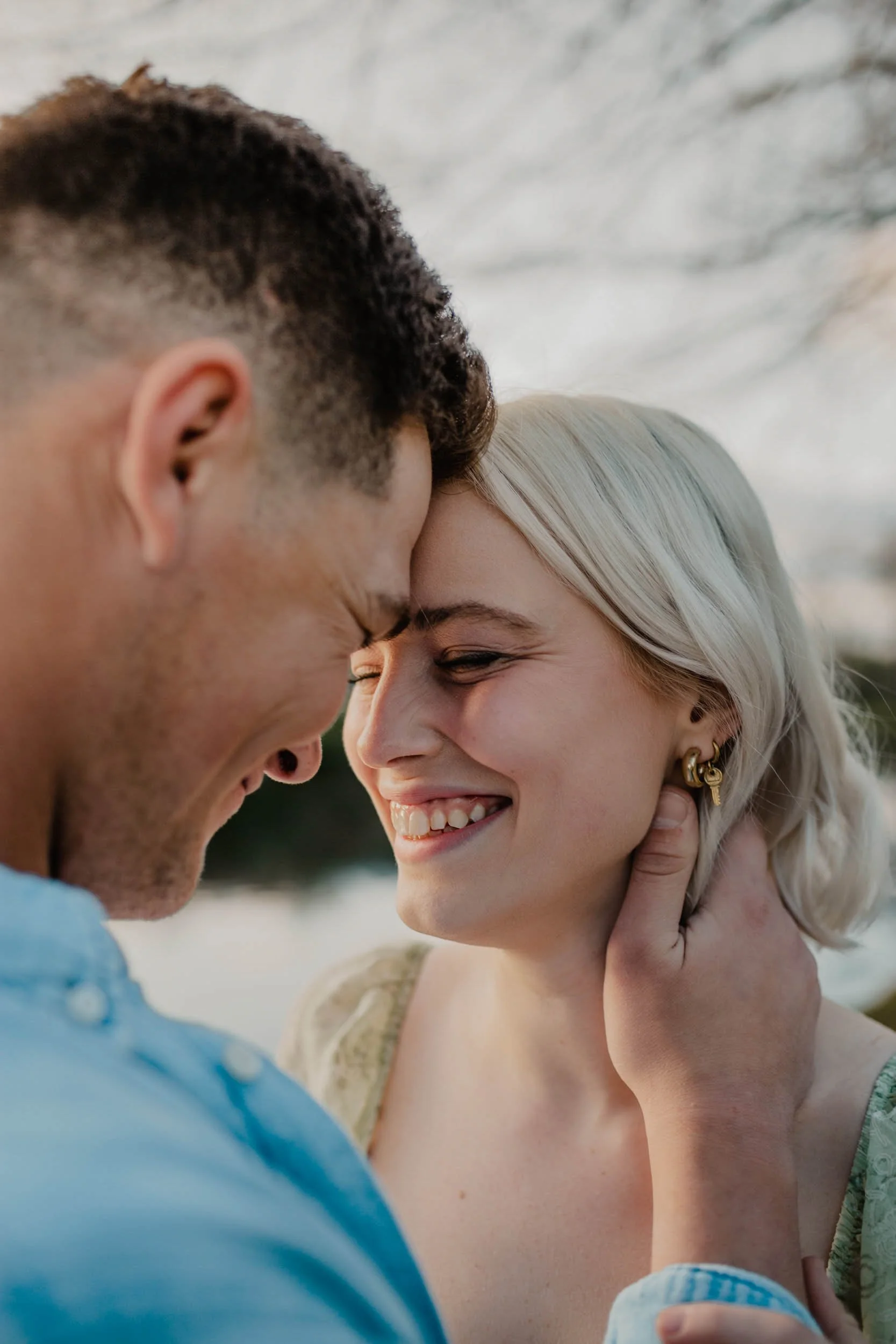 A man and a woman are smiling and touching foreheads closely, outdoors during the day.