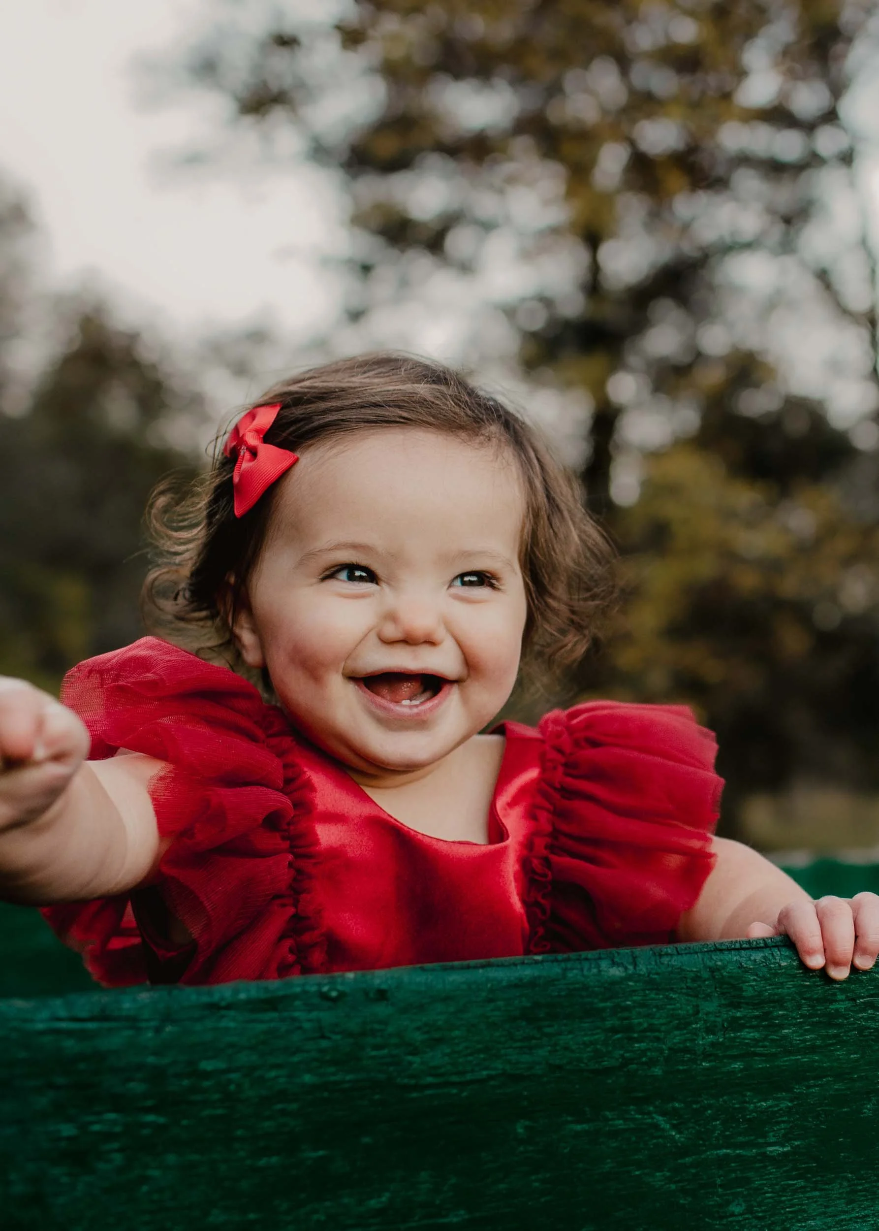 A young girl with brown curly hair, wearing a red dress with puffy sleeves and a red bow in her hair, smiling and reaching out from behind a green wooden structure outdoors with trees in the background.