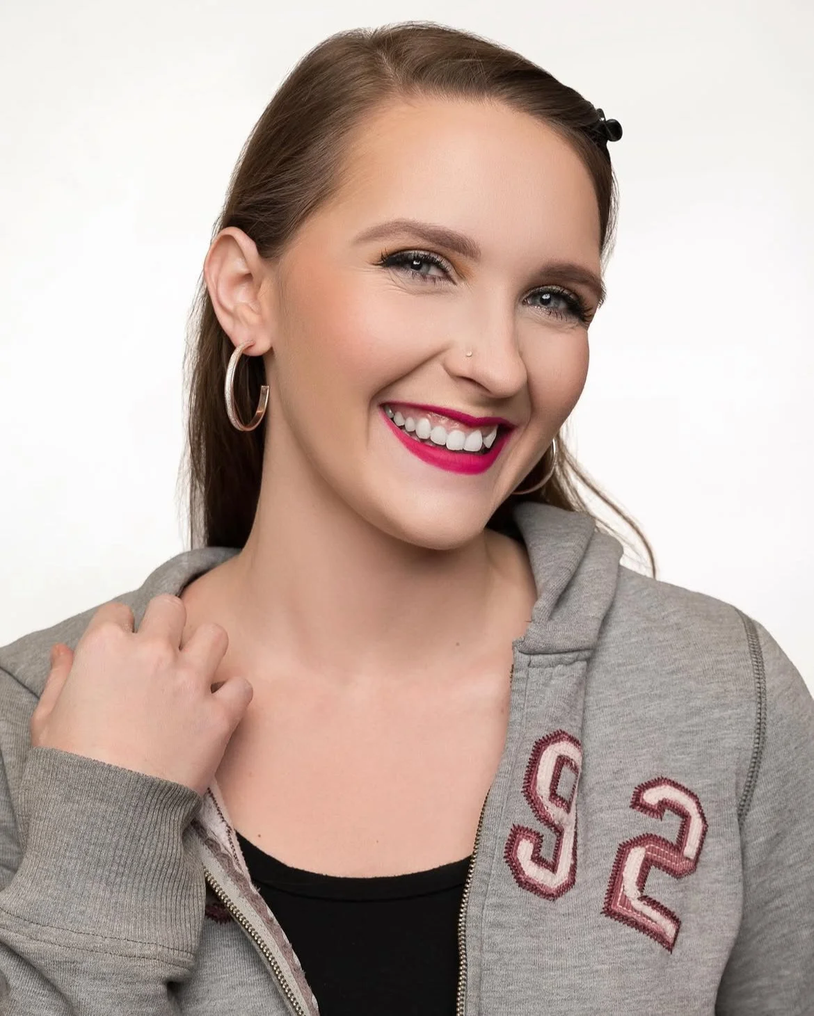 A young woman with long brown hair, wearing hoop earrings, a gray zip-up hoodie, and pink lipstick, smiling and looking at the camera.