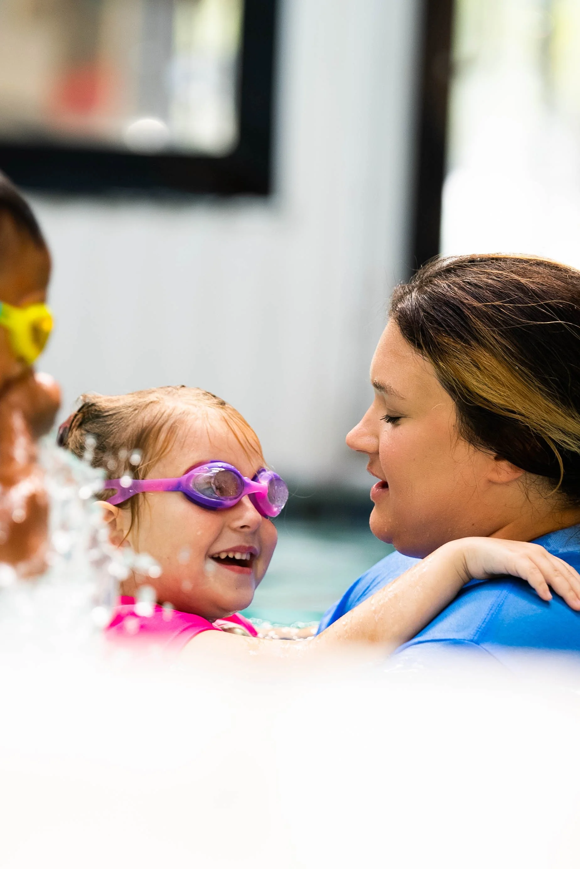 A young girl with red hair and purple goggles is smiling at an adult woman in a swimming pool. The girl and woman are facing each other, with the girl having her arms around the woman's shoulders. There are other children with swim goggles in the background.