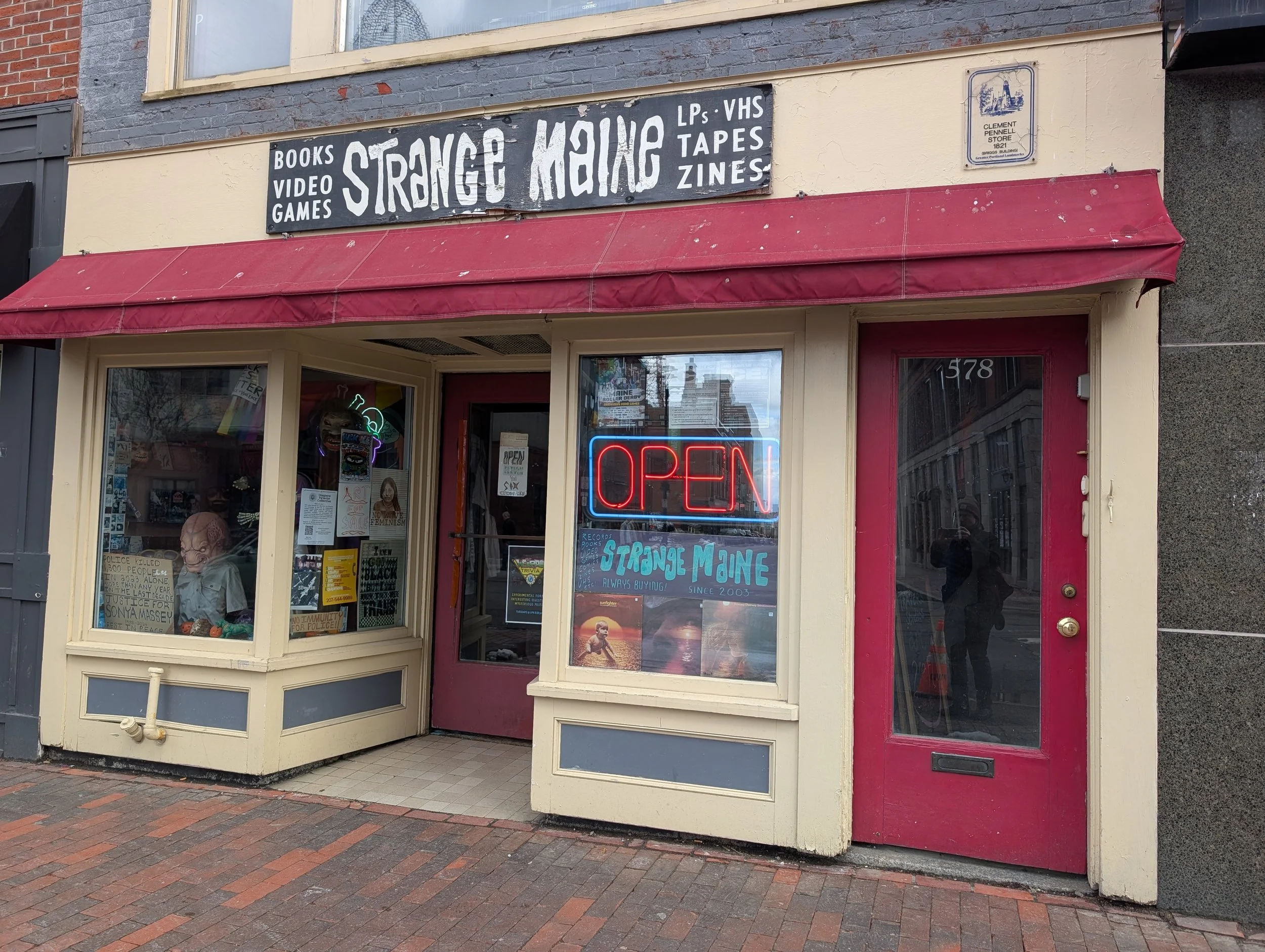 Wooden storefront with a narrow signboard above and a narrow glass door flanked by glass windows covered on the inside with flyers and merchandise