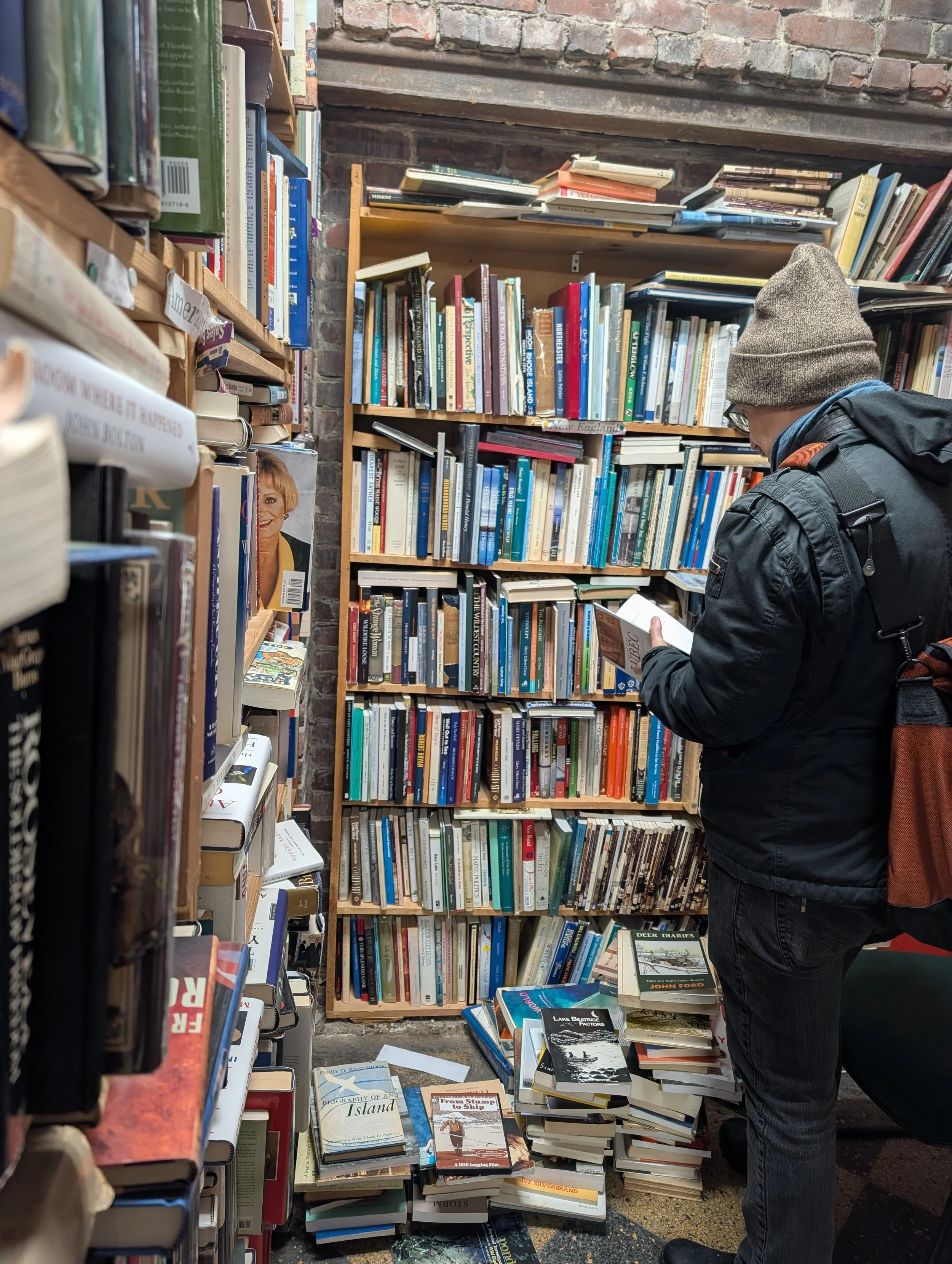 A white man perusing a book in a corner defined by shelves teeming with books stacked in several ways