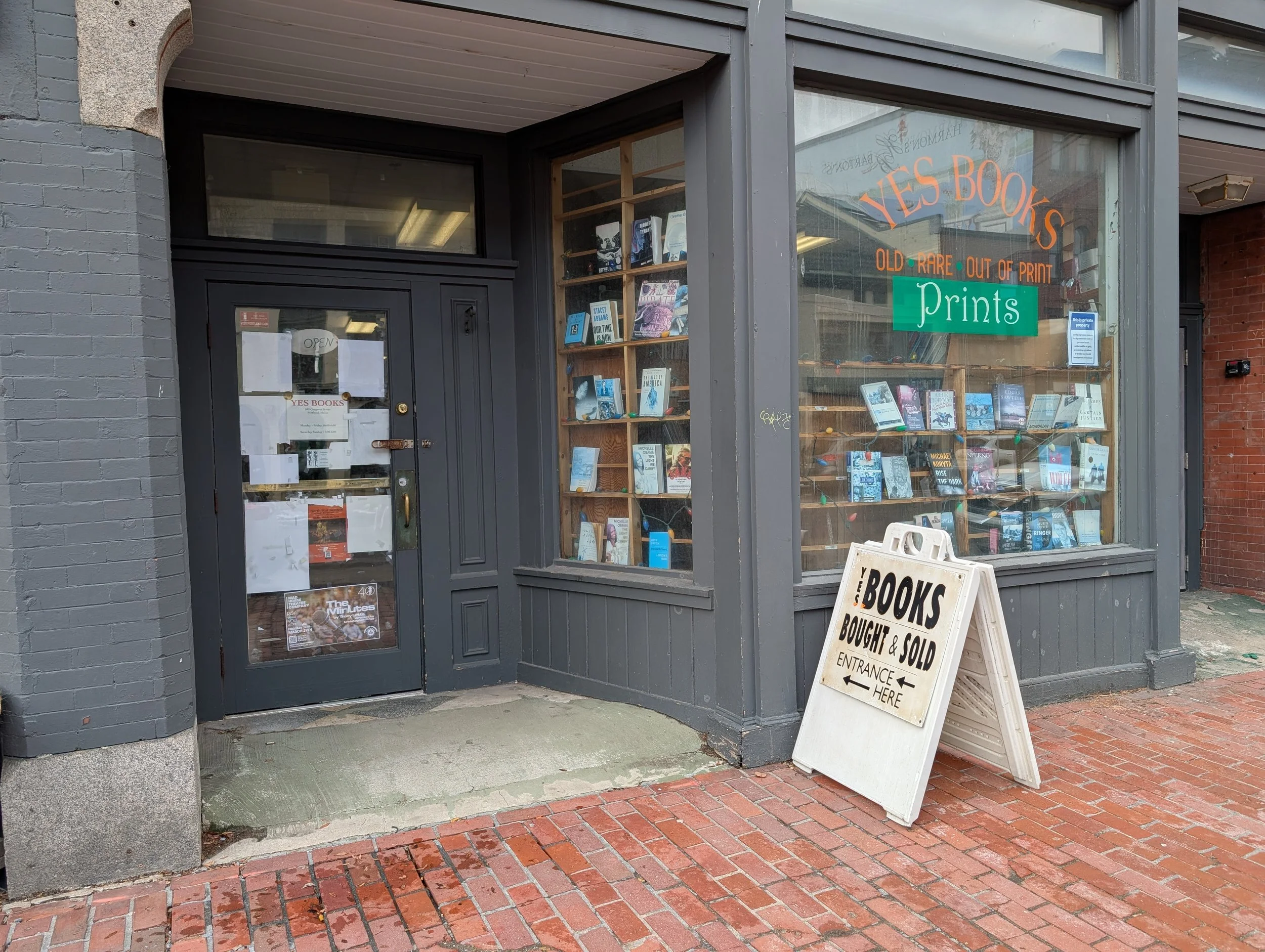 Brick and wood storefront with company logo and books in the window and an a-frame sign out front on the brick sidewalk