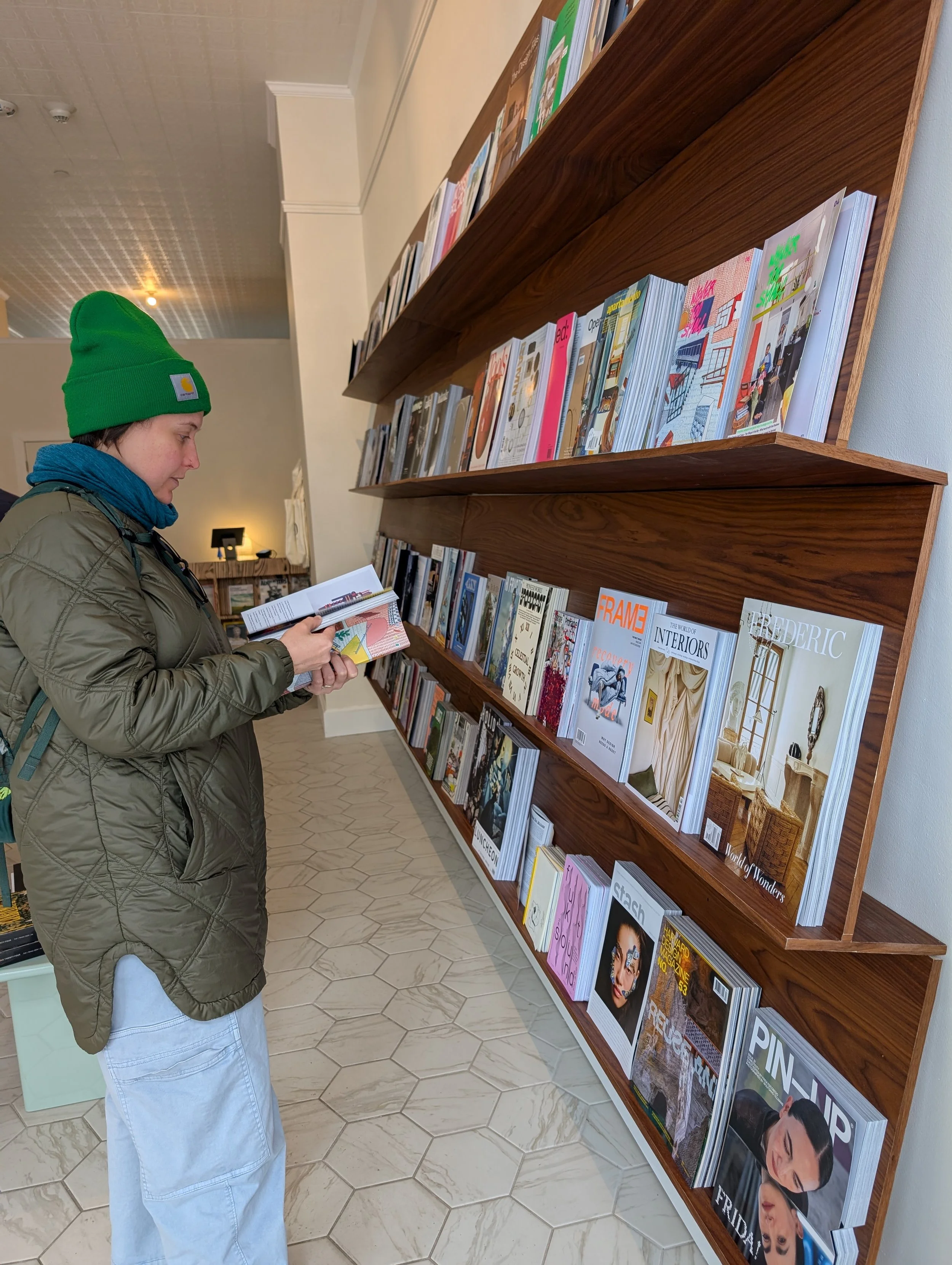 A white woman in winter coat and hat perusing a magazine in front of a large wooden shelf fixed to the wall and displaying many more magazines