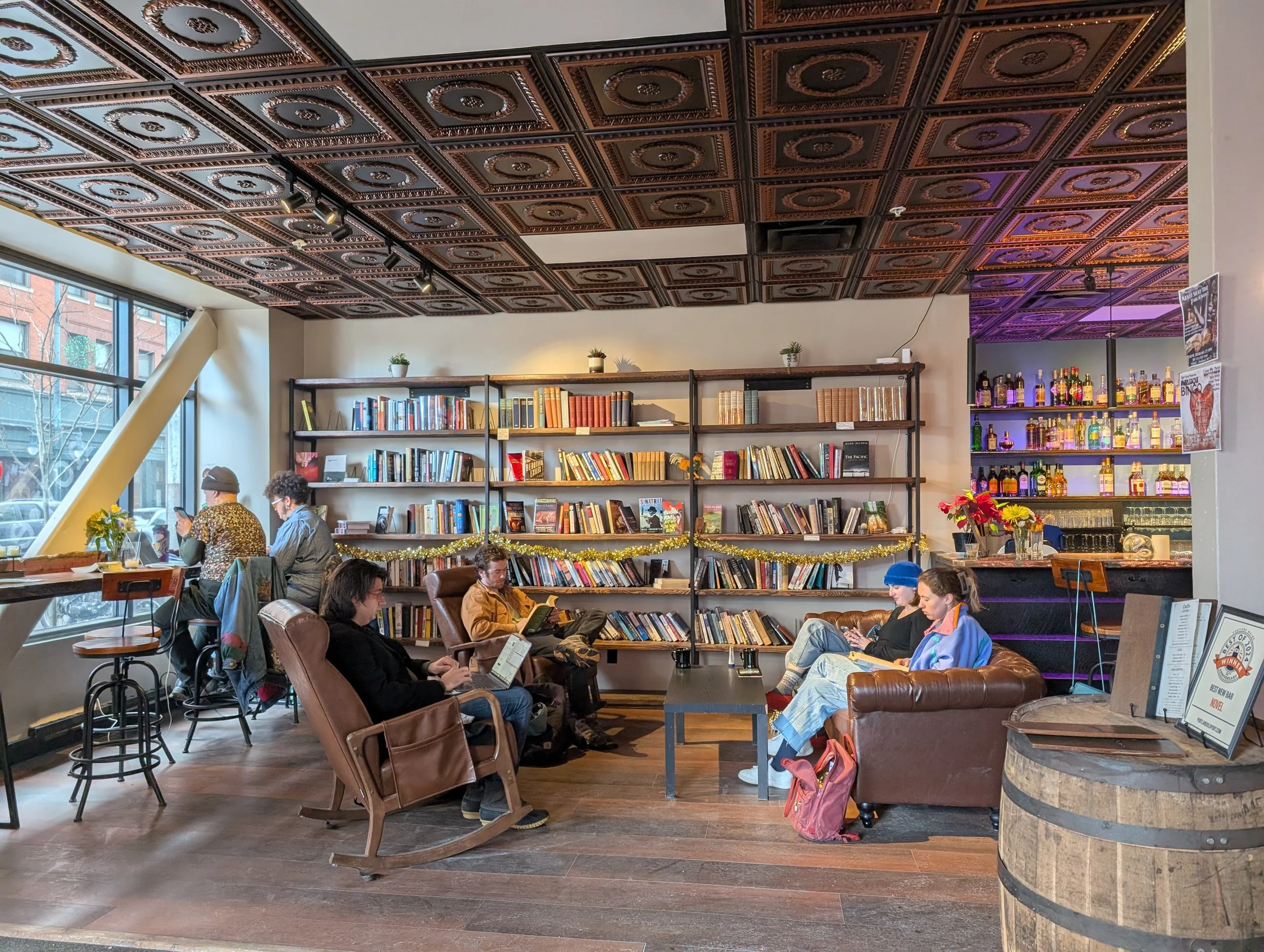 Sun-dappled space with wooden floor, seating set up like a living room with people reading and working on laptops with a stocked bookshelf on the wall behind them