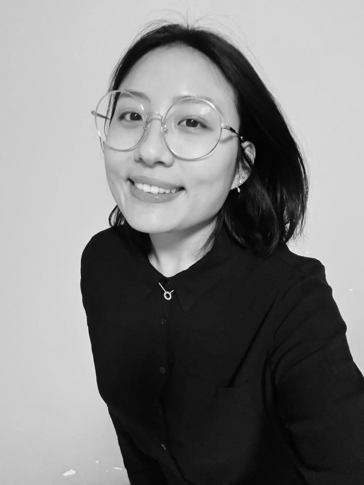 A smiling woman with glasses and earrings, wearing a dark collared shirt, takes a selfie against a plain background.
