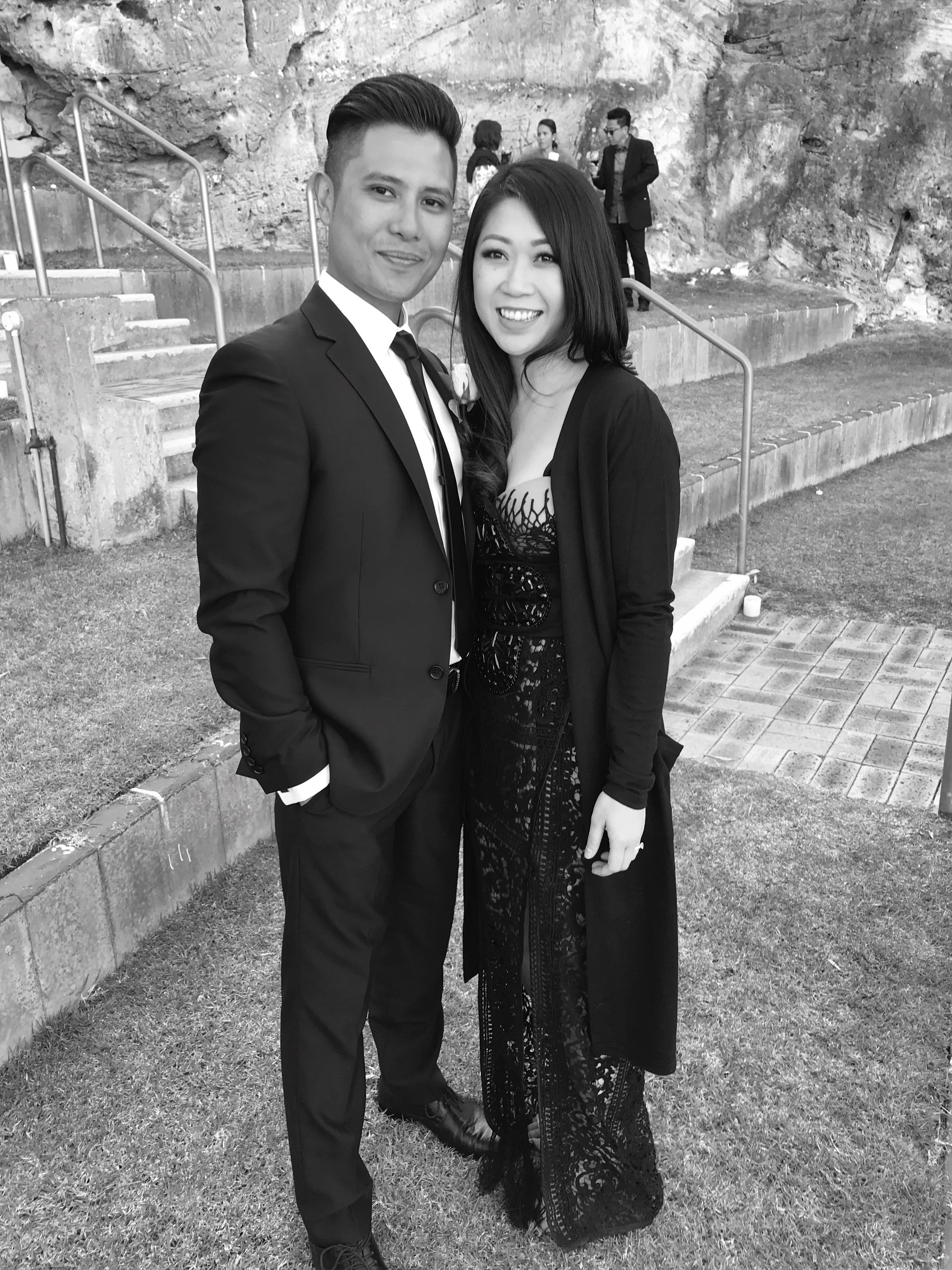 A young couple dressed in formal attire, standing outdoors on a grassy area with stairs and a rocky wall in the background. The man wears a suit with a tie, and the woman wears a lace dress and cardigan, both smiling at the camera.