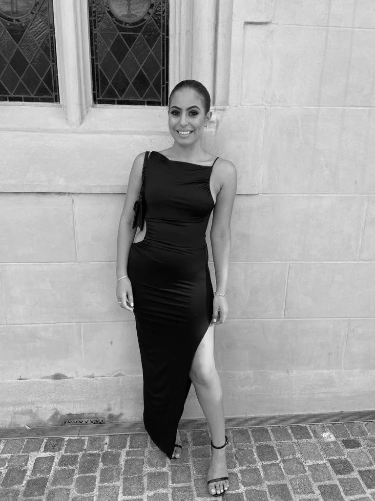 A woman in a black evening gown with a thigh-high slit, standing outdoors on cobblestone pavement in front of a stone wall with windows.