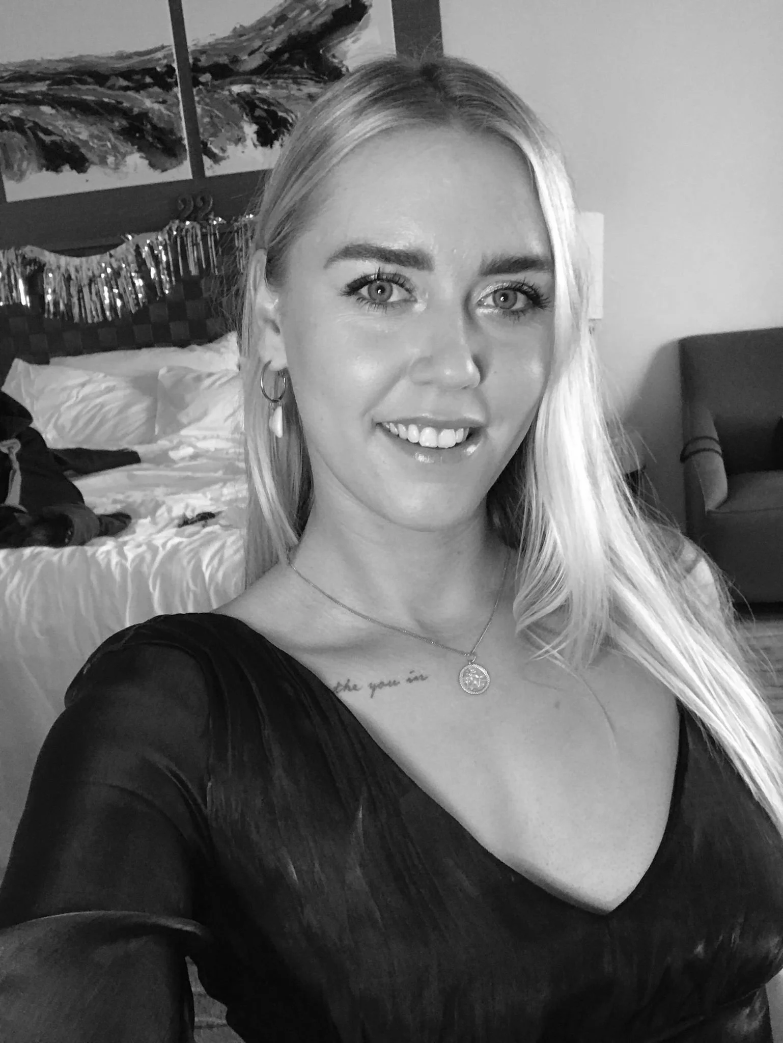 A black and white photo of a woman with long blonde hair smiling and taking a selfie in a room.