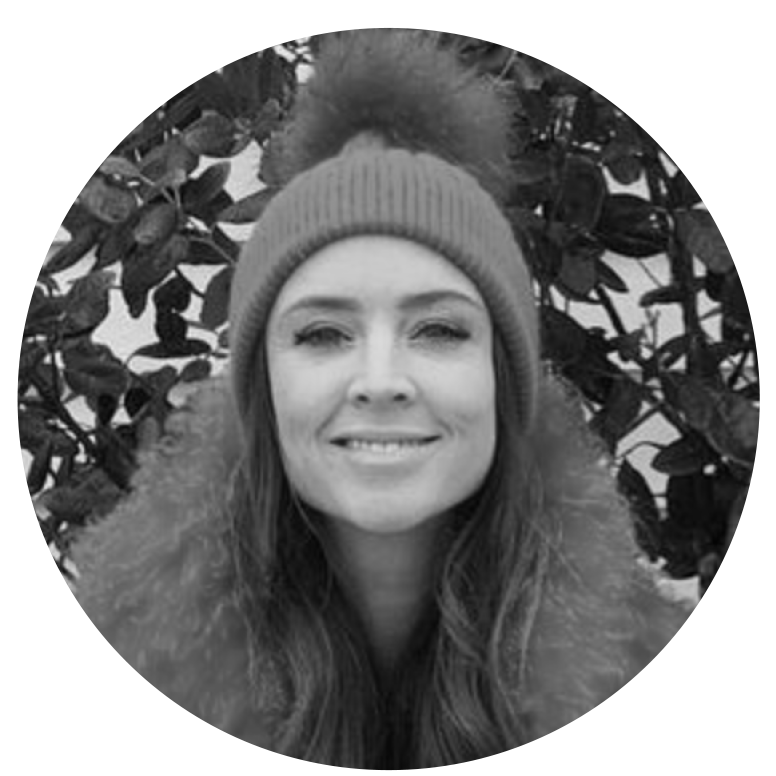 A woman in a beanie hat with a pom-pom, smiling and standing outdoors in front of leafy bushes, in black and white.