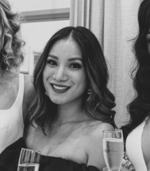 Young woman with shoulder-length wavy hair smiling at a social gathering, holding a glass of what appears to be champagne, in a black-and-white photo.