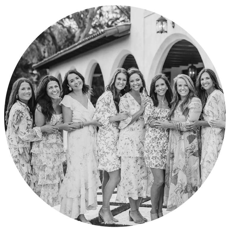 A group of eight women in dresses, standing outdoors in front of a building with arches, smiling and holding each other.