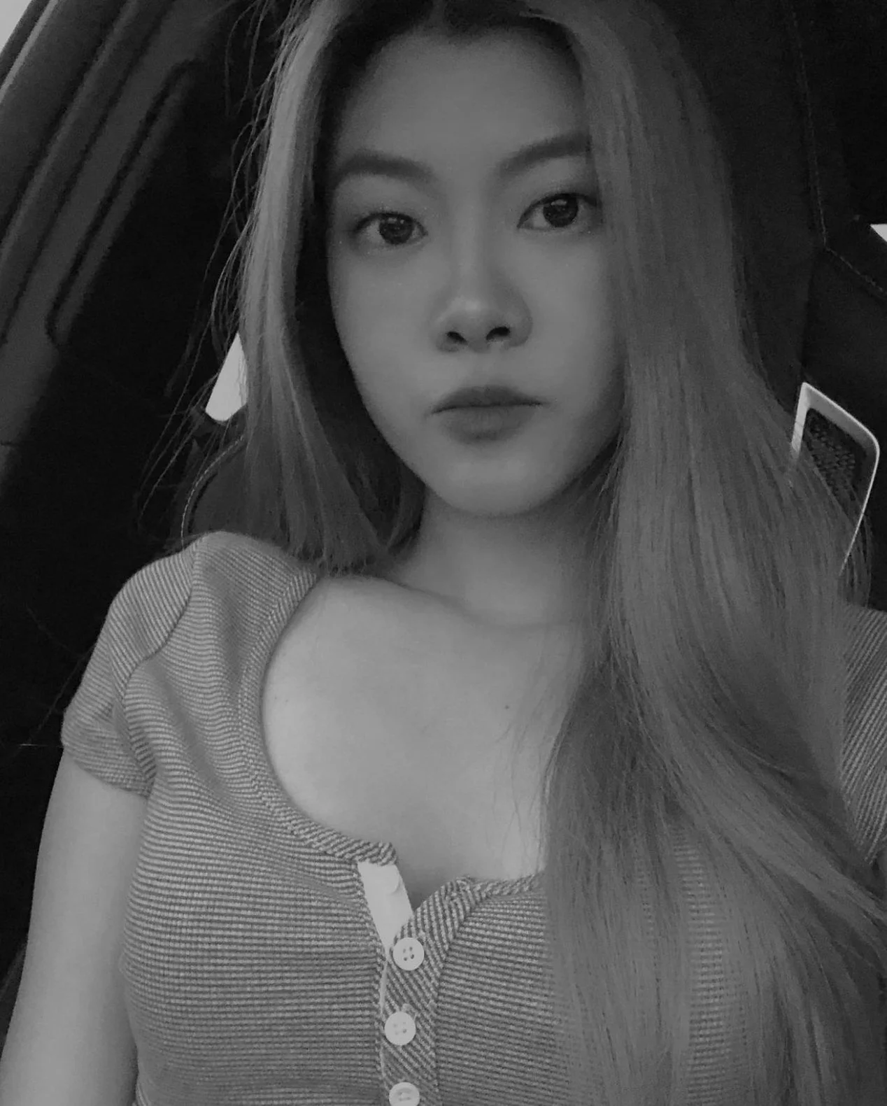 A black and white close-up photograph of a young woman with long straight hair, wearing a buttoned top, taking a selfie.