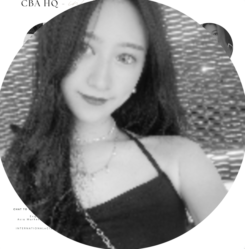 Black and white photo of a woman with long dark hair, wearing a sleeveless top and a necklace, sitting on a woven chair, smiling at the camera.