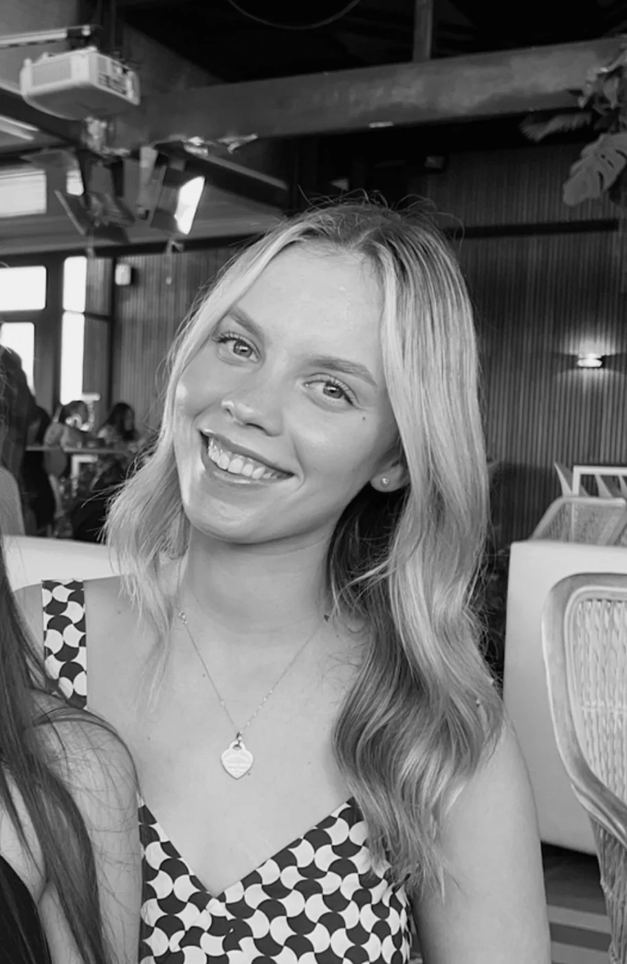 A smiling young woman with wavy hair, wearing a polka dot dress and a necklace, sitting indoors at a social gathering.