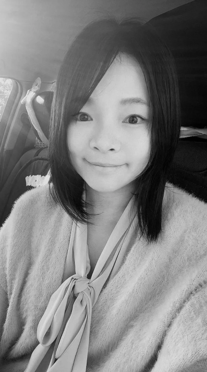 A woman with shoulder-length dark hair smiling while sitting in a car, wearing a light-colored sweater and a neck scarf.