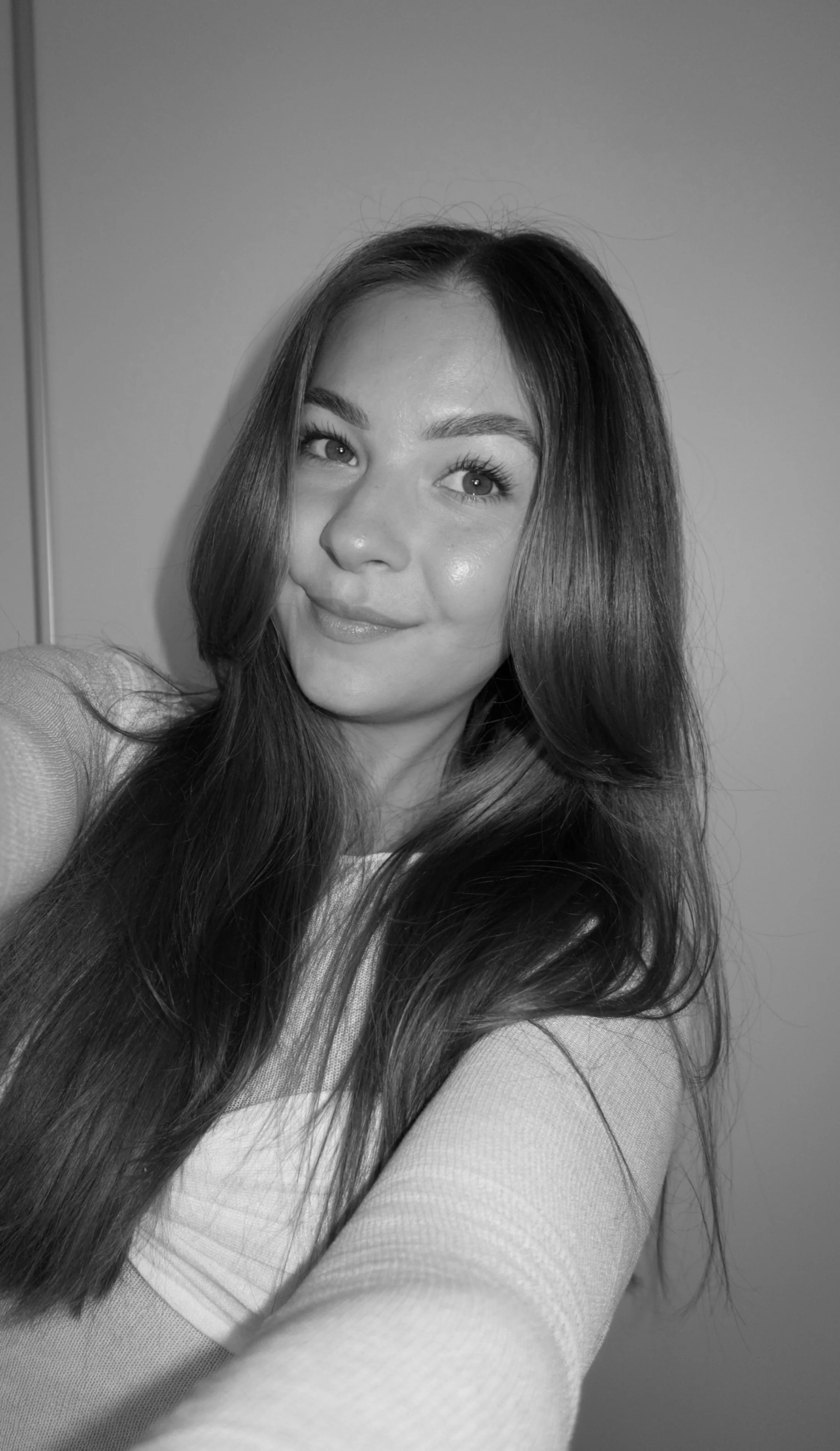 A young woman with long hair taking a selfie, smiling slightly, in black and white.