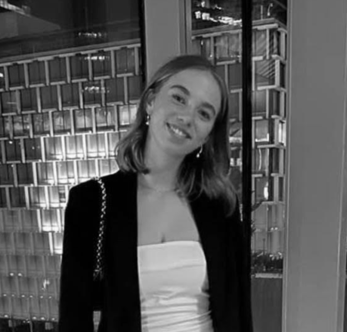 A woman with shoulder-length hair, wearing a strapless top and a black blazer, smiling and posing indoors near a window with a cityscape view at night.